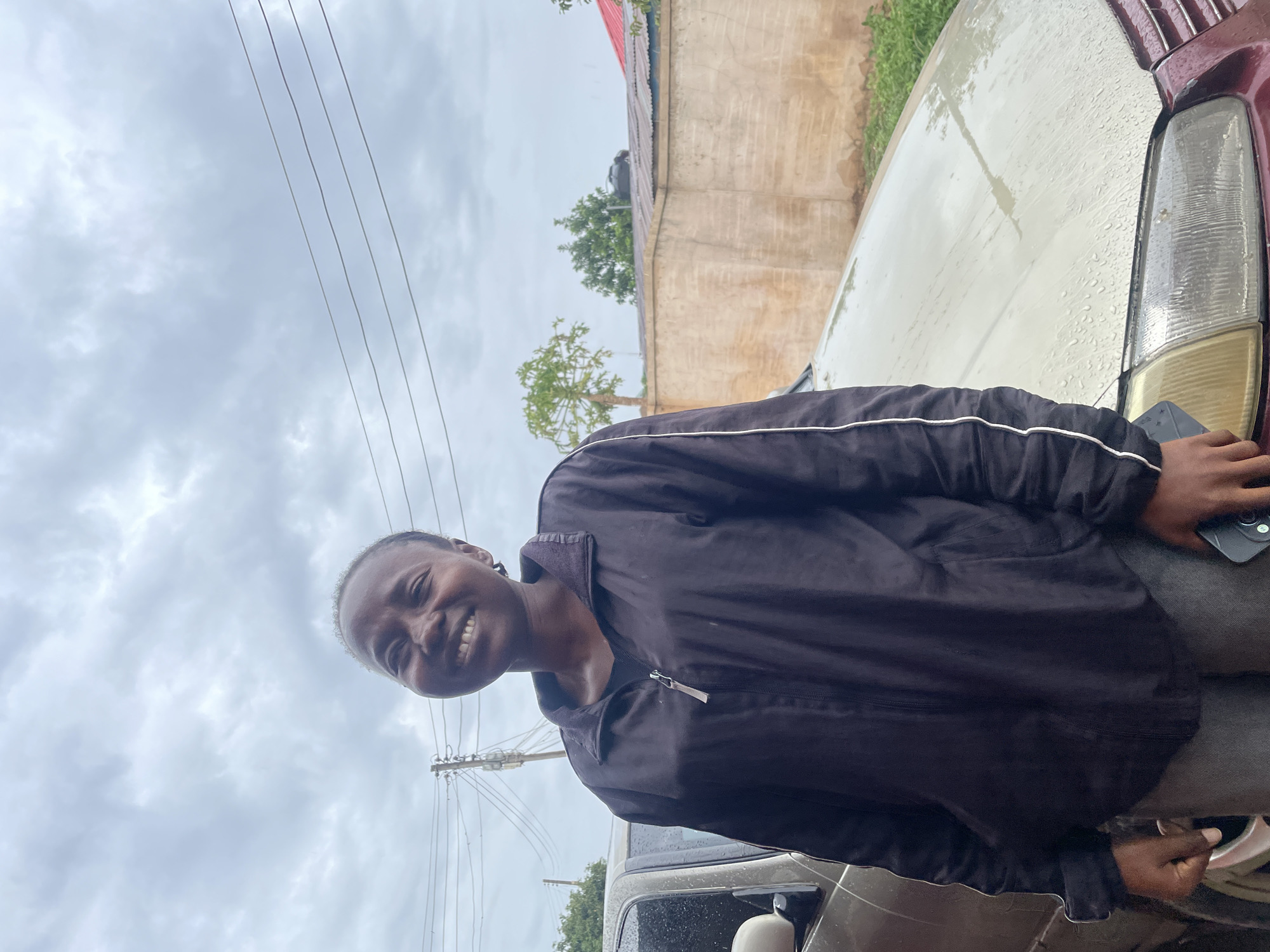 A smiling young woman is seen in Jos, Nigeria. The scene appears to be outdoors on a cloudy day. She is wearing a black jacket with white trim. Adjacent to her are parts of two vehicles, one maroon and one grey. A building is visible behind her, and there are utility poles and wires in the background. Raindrops are visible on the surface of the car. She is holding a black smartphone in her right hand.