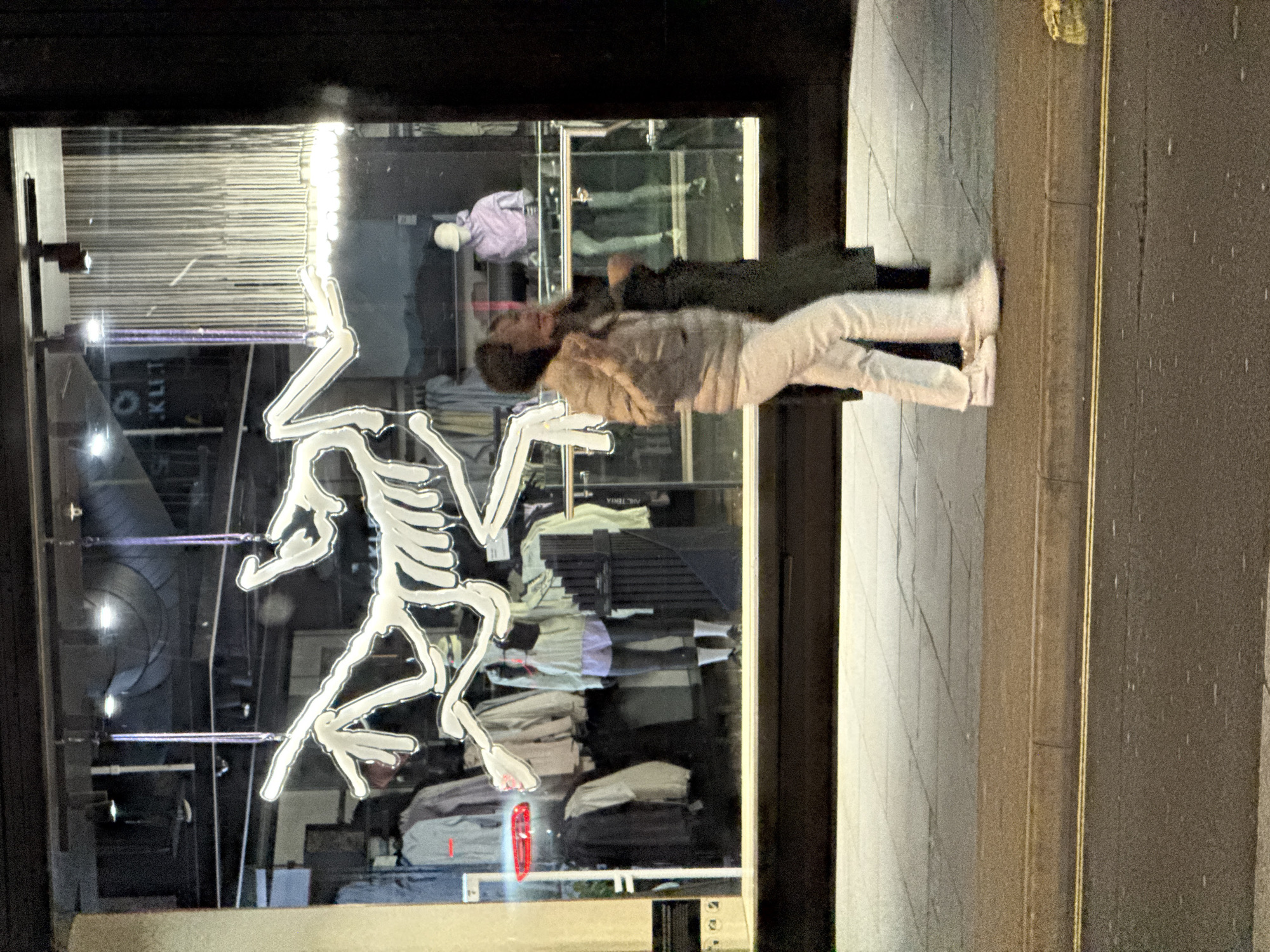 An upright-oriented, dark, and somewhat grainy night-time photograph captures a street scene outside a retail store in London. The dominant feature is a large storefront window illuminated from within, displaying merchandise and a prominent neon skeleton outline applied to its exterior.
A person is captured in motion, walking past the store window from left to right. They appear to be wearing light-colored trousers and a patterned or textured top. Their reflection is clearly visible in the window, overlapping with the store's interior and the neon skeleton. The person's face is somewhat obscured by motion blur and the angle.
Inside the brightly lit store, several mannequins are visible, dressed in clothing such as a purple shirt and dark pants. Racks of clothing can be seen further inside. The window also reflects the street or pavement outside, with hints of yellow lines that could be road markings. A red light, possibly a car's tail light or a reflection of a distant sign, is visible within the store's reflection.
On the store window, in addition to the neon skeleton, parts of text are discernible, including what appears to be "KLIT" (likely part of a larger brand name like "ARC'TERYX", of which "ARCTERIA" is also faintly visible) on a sign within the store. The overall atmosphere is urban and nocturnal, with the store acting as a beacon of light against the dark surroundings.