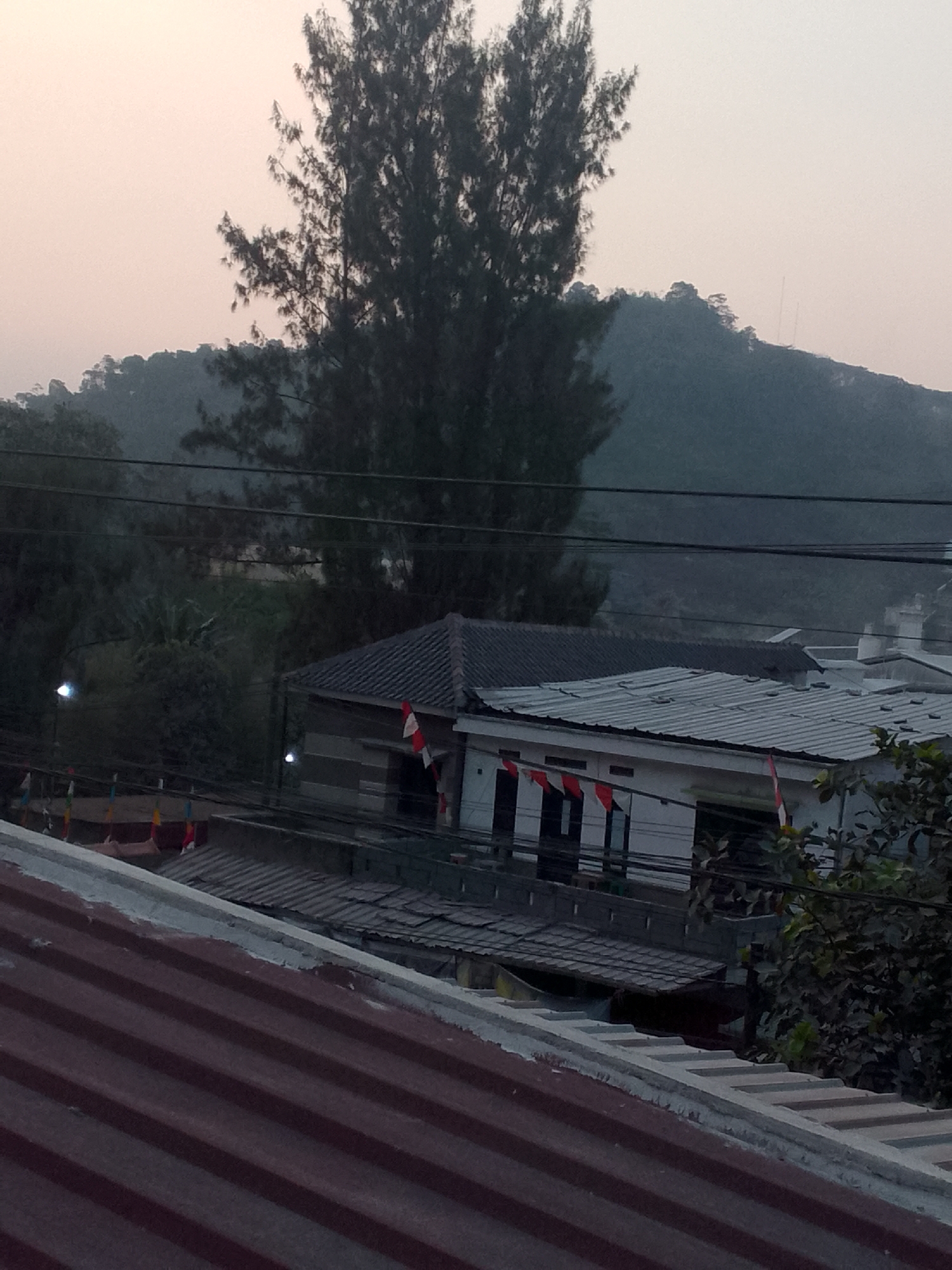 The image depicts an outdoor scene, likely in the city of Gunungmasigit, Indonesia, as indicated by the presence of Indonesian flags. The foreground is dominated by a reddish-brown corrugated metal roof. Behind this, several other buildings are visible, characterized by various roofing materials, including gray corrugated metal and what appears to be tile. One building in the mid-ground has white walls with dark-framed windows and features multiple Indonesian flags, red and white, displayed along its facade. Another building to the left has a dark, possibly tiled roof. The environment includes lush green foliage surrounding the structures, and a large tree with dark, dense foliage stands prominently in the mid-ground. In the background, a hazy, tree-covered hill rises, with what appear to be radio antennas on its crest. The sky is overcast with a pale, diffused light, suggesting either early morning or late afternoon. Power lines crisscross the scene, indicating an inhabited area. No people are visible in the image.
