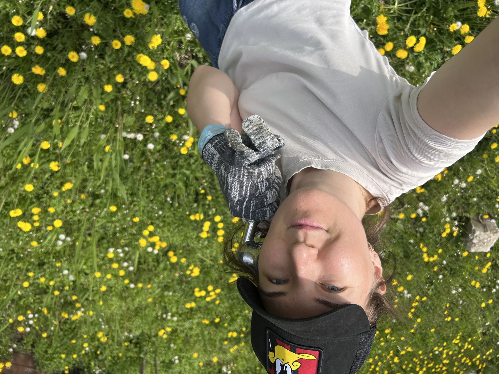 A medium shot depicts a person lying on a grassy field covered with dandelions and other small white flowers. The person has fair skin, blue eyes, and is wearing a black baseball cap with a colorful cartoon patch, a light-colored t-shirt, and blue jeans. They are holding a gardening tool with a grey and black striped glove on their hand. The location is potentially near the city of Noviy, Russia, based on surrounding vegetation.