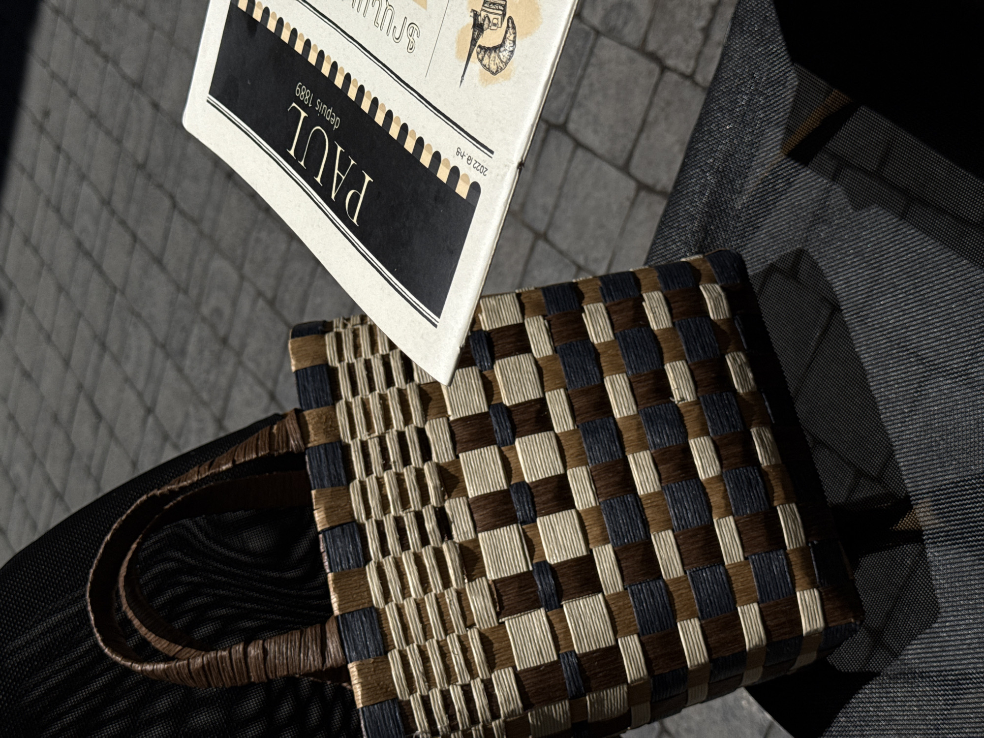 A woven tote bag with brown, beige, and dark blue squares sits on a dark-colored fabric surface, possibly a chair or bag. To the right of the bag, a menu or brochure with the logo "PAUL depuis 1889" is partially visible, laid out on the same surface. The background shows a paved outdoor area with cobblestone-like patterns, illuminated by sunlight casting distinct shadows. The scene appears to be outdoors, likely in a cafe or public seating area. No people are visible in the frame. The lighting suggests a daytime setting with strong directional sunlight. The overall composition is a close-up shot focusing on the bag and the menu against a textured ground. The image does not provide any specific visual cues to indicate a location in Yerevan, Armenia.