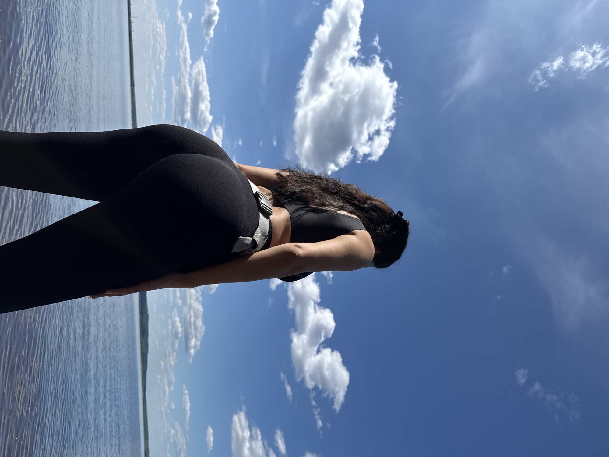 A person, appearing female, is depicted from a low-angle perspective, viewed from the back and slight right profile, standing on the edge of a large body of water. She wears a black sports bra or crop top and black form-fitting leggings. A light grey or white utility belt with a silver-colored buckle is worn around her waist. Her dark, long, wavy hair is pulled back, secured with a dark hair clip visible at the top of her head. She stands with her weight slightly shifted, looking towards the right side of the frame, facing the water and the distant horizon. The upper two-thirds of the image are dominated by a clear blue sky with scattered white cumulus clouds, indicating a bright, sunny day. The lower third features the calm, expansive surface of the Votkinsk Reservoir, with subtle ripples visible across the water. A faint, distant landmass, likely a treeline or low hills, is visible along the horizon line. The scene takes place outdoors in the vicinity of Chaikovsky, Russia, on the Votkinsk Reservoir.