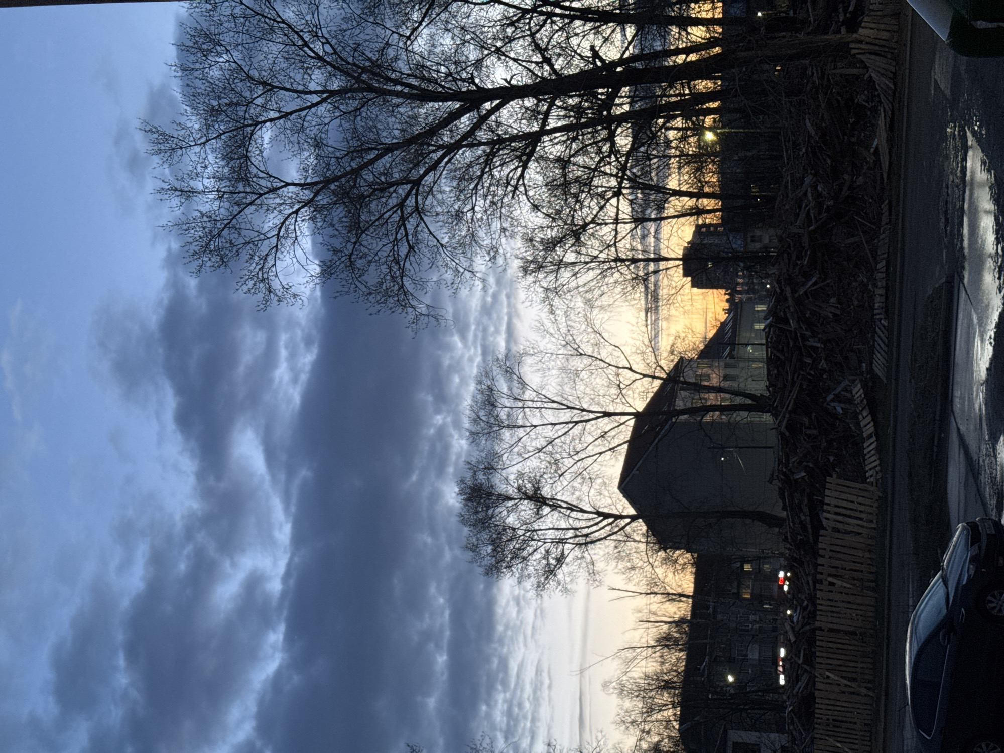 This landscape image, taken in Chaikovsky, Russia, depicts a scene at either dawn or dusk, characterized by a dramatic sky and silhouetted urban elements.
The sky dominates the upper portion of the frame, presenting a striking contrast. On the left, heavy, dark blue-grey clouds create a somber mood, gradually transitioning towards the right into lighter, more wispy clouds. The far right side of the sky glows with warm hues of yellow and orange, indicating the low position of the sun.
Numerous bare-branched trees, suggestive of late autumn or winter, are silhouetted against the sky, their intricate network of branches framing the scene from the top and bottom.
In the midground, several dark buildings are partially visible, mostly in silhouette, with a distinct roofline of one building standing out. A large, prominent pile of wooden pallets or discarded lumber forms a significant barrier across the mid-right. Faint lights from within buildings or distant streetlights are discernable in the darker areas.
The foreground on the far right features a wet road or pavement with a large puddle reflecting the light, and a dark passenger car is parked alongside it. The overall atmosphere is stark and moody, capturing the transitional light between day and night in an urban or semi-urban environment. No visible text is present in the image.