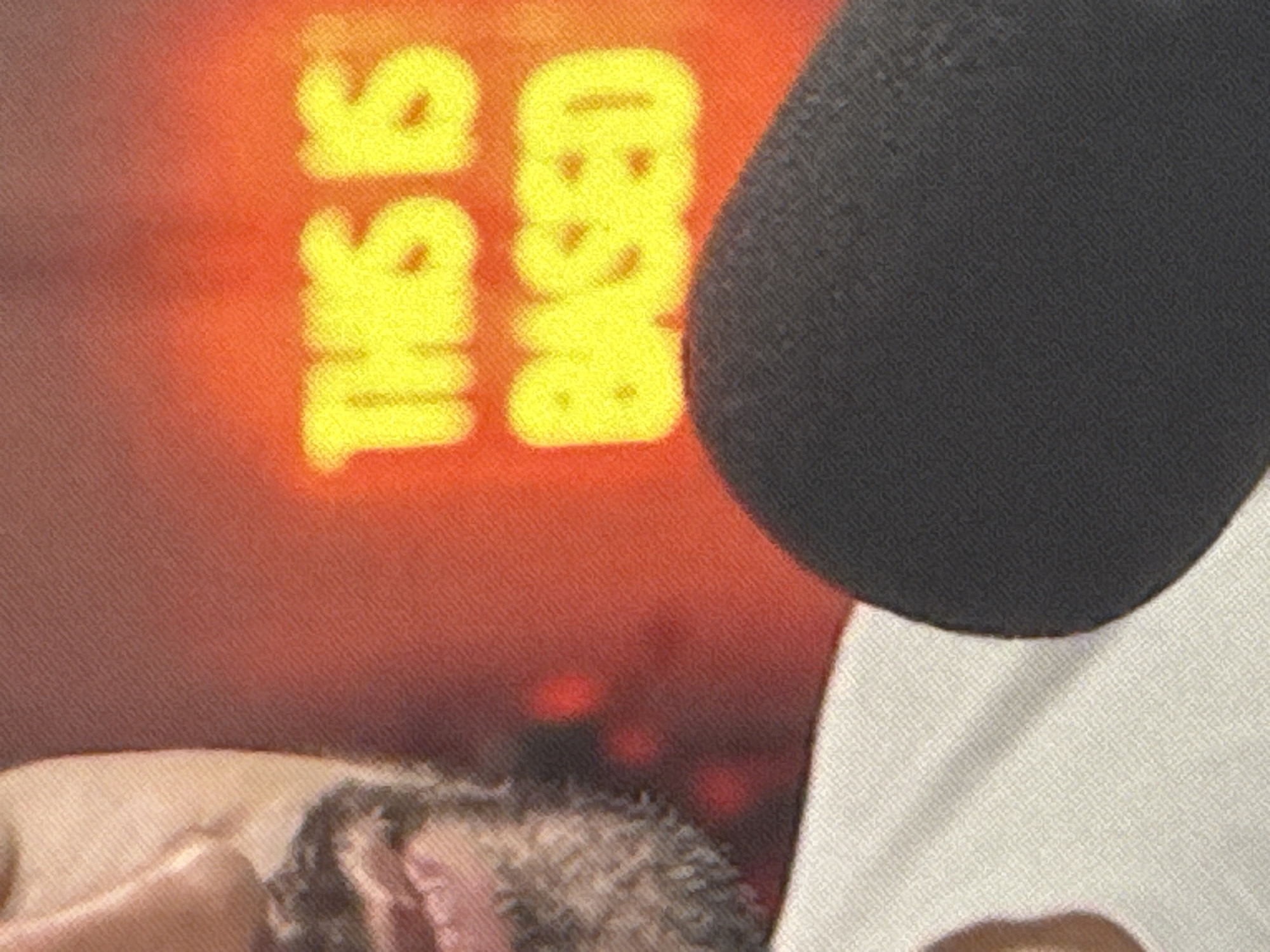 The image shows a close-up of a person's face and a microphone. The person has a beard and is wearing a white shirt. Behind the person is a neon sign that says "THIS IS BASED" in yellow lettering. The sign is on a red and orange gradient background. The image does not provide any information about the location in the city of Saratov, Russia.