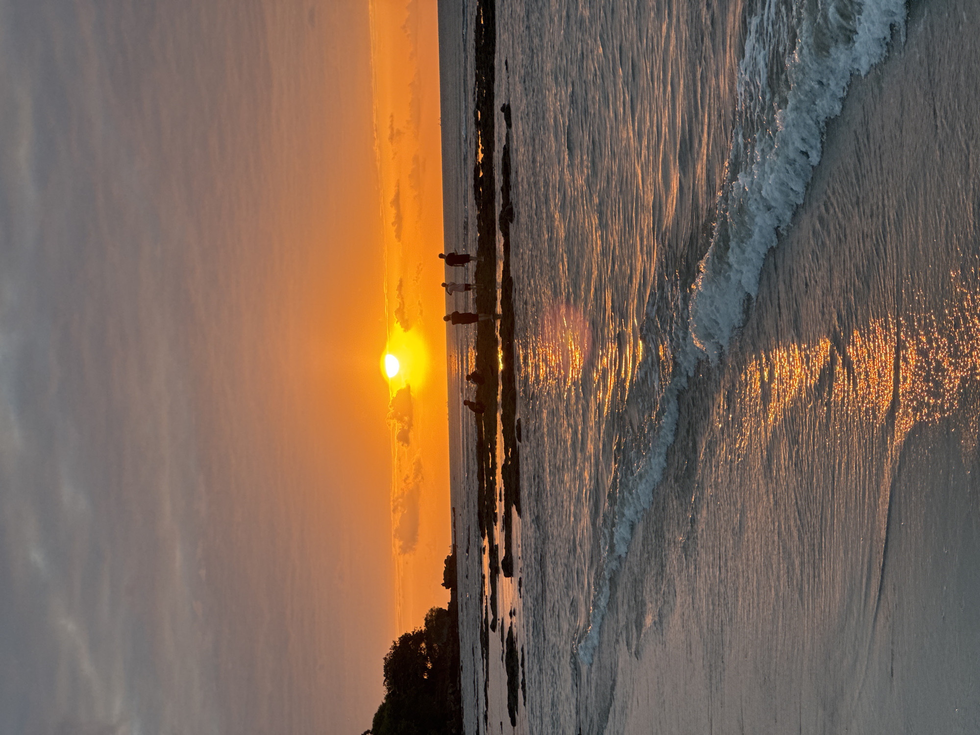 The image depicts a serene beach scene in Kuta Selatan, Indonesia, during sunset. The sky is a dramatic gradient of orange and grey clouds, with the sun prominently positioned as a bright, glowing orb near the horizon. The sun's reflection shimmers on the water's surface, creating a path of golden light that stretches towards the shore.
Several individuals are visible standing on rocky outcrops in the shallow water, silhouetted against the vibrant sunset. Some are gathered in small groups, while others appear to be standing alone. The waves gently lap against the sandy beach in the foreground, with a few whitecaps visible. The overall atmosphere is tranquil and picturesque, capturing the natural beauty of a tropical sunset.
