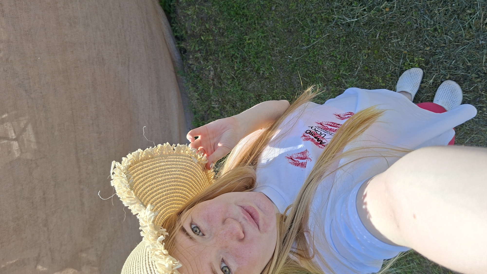 A woman with long red-blonde hair, wearing a straw hat with a fringed edge, a white t-shirt with red lip-print graphics and white open-toe sandals, is photographed from a high angle. She is lying on a grassy surface. A brown woven fabric or curtain is visible in the upper-left corner of the frame. The location is outdoors, near Большой Букор, Russia.
