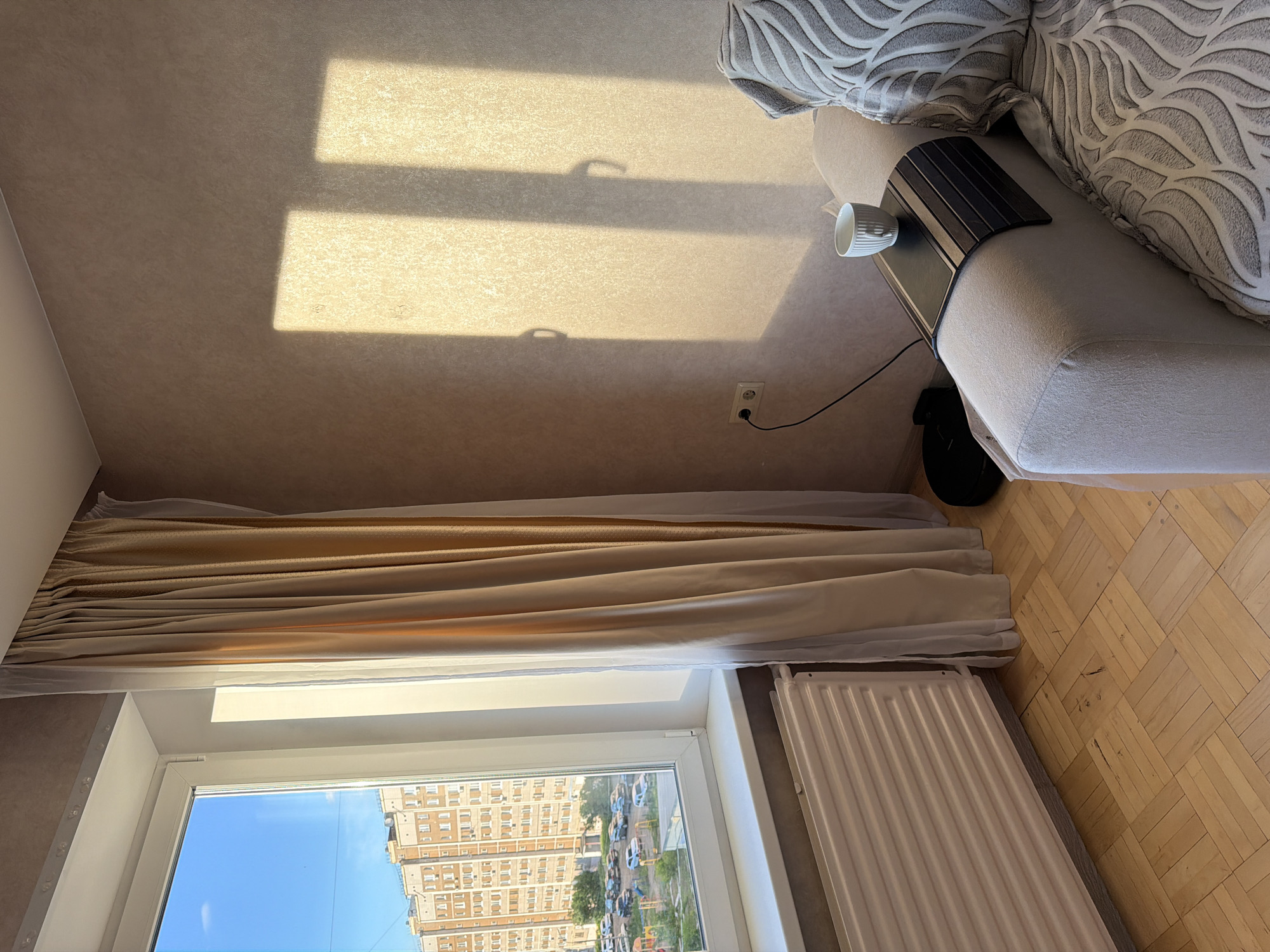 The image depicts an interior scene in Izhevsk, Russia, observed through a window during daylight. Outside, a multi-story residential building is visible under a clear blue sky. The scene is viewed from an elevated position, possibly within an apartment. Inside, a sofa is present with a cup and a small side table attached to the armrest. Curtains partially obscure the window, and a radiator is positioned near the window frame. The room has a light-colored textured wall and a parquet wood floor. There are no people visible in the image.