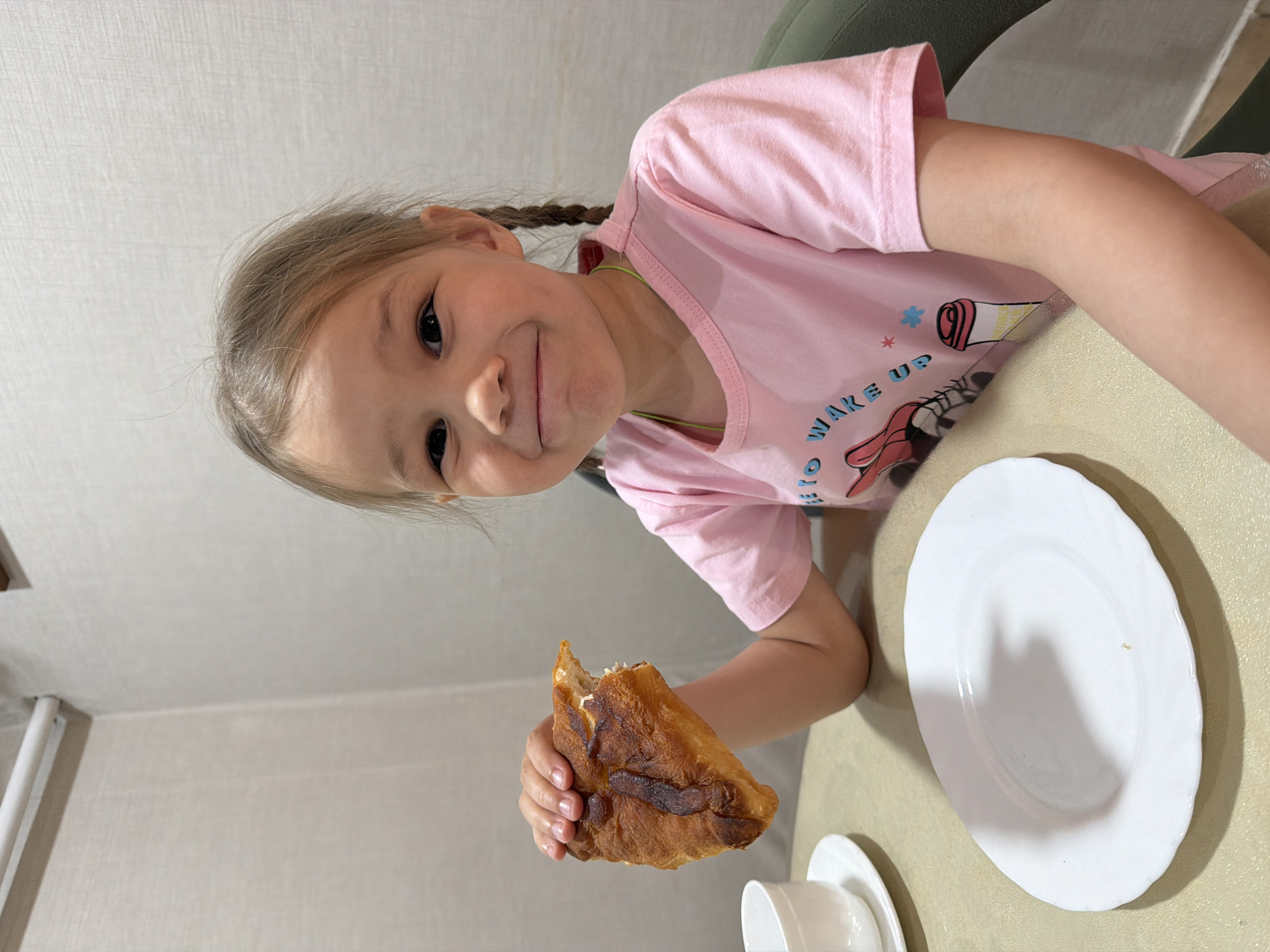 A young girl with braided hair, wearing a pink t-shirt, is at a table with a white plate, a white cup, and eating a fried bread snack. The location is Chaikovsky, Russia.