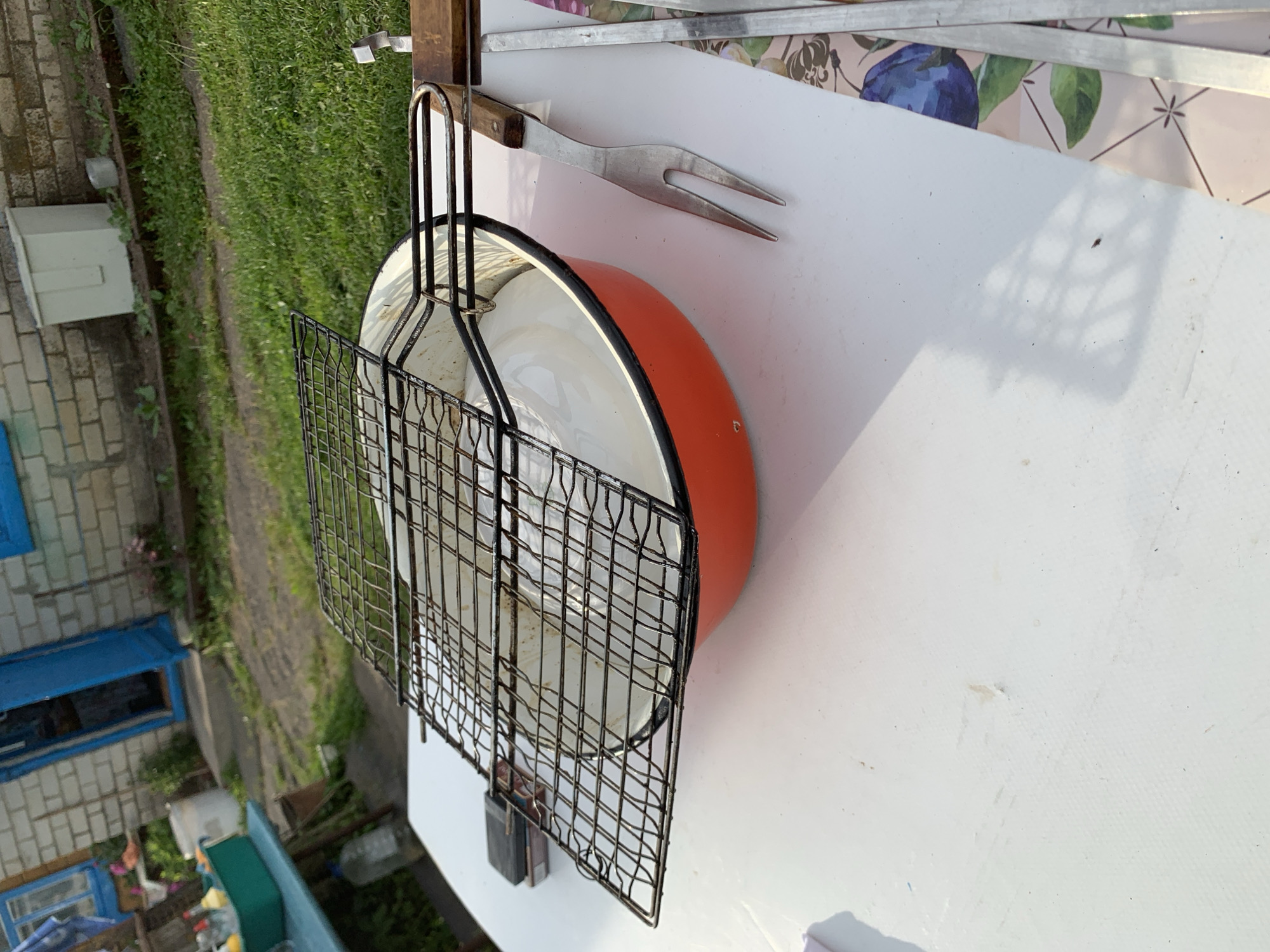 The image depicts an outdoor setting in Відзаўскі сельскі Савет, Belarus. In the foreground, a white table surface is visible, upon which a rectangular metal grill grate, showing signs of use and residue, rests over an orange enamel basin with a white interior. To the right of the basin, a two-pronged metal grilling fork with a wooden handle is positioned. A red and black matchbox is located on the table to the left of the grill grate. A patterned mat with floral and fruit designs is partially visible on the far right edge of the table. Sunlight illuminates the scene, casting distinct shadows on the table.
In the midground, to the left, a light blue table holds a blue-green rectangular container containing various objects. A large clear plastic bottle is on the ground beneath this table. The ground is covered with green grass.
The background features a brick building, constructed with light-colored bricks. Two blue-framed windows are visible on the building, along with a blue-framed doorway, which appears partially open. Sparse vegetation grows near the base of the building. A white, rectangular utility box is affixed to the brick wall on the right side of the frame. A white bucket is present on the ground near the blue table on the left.