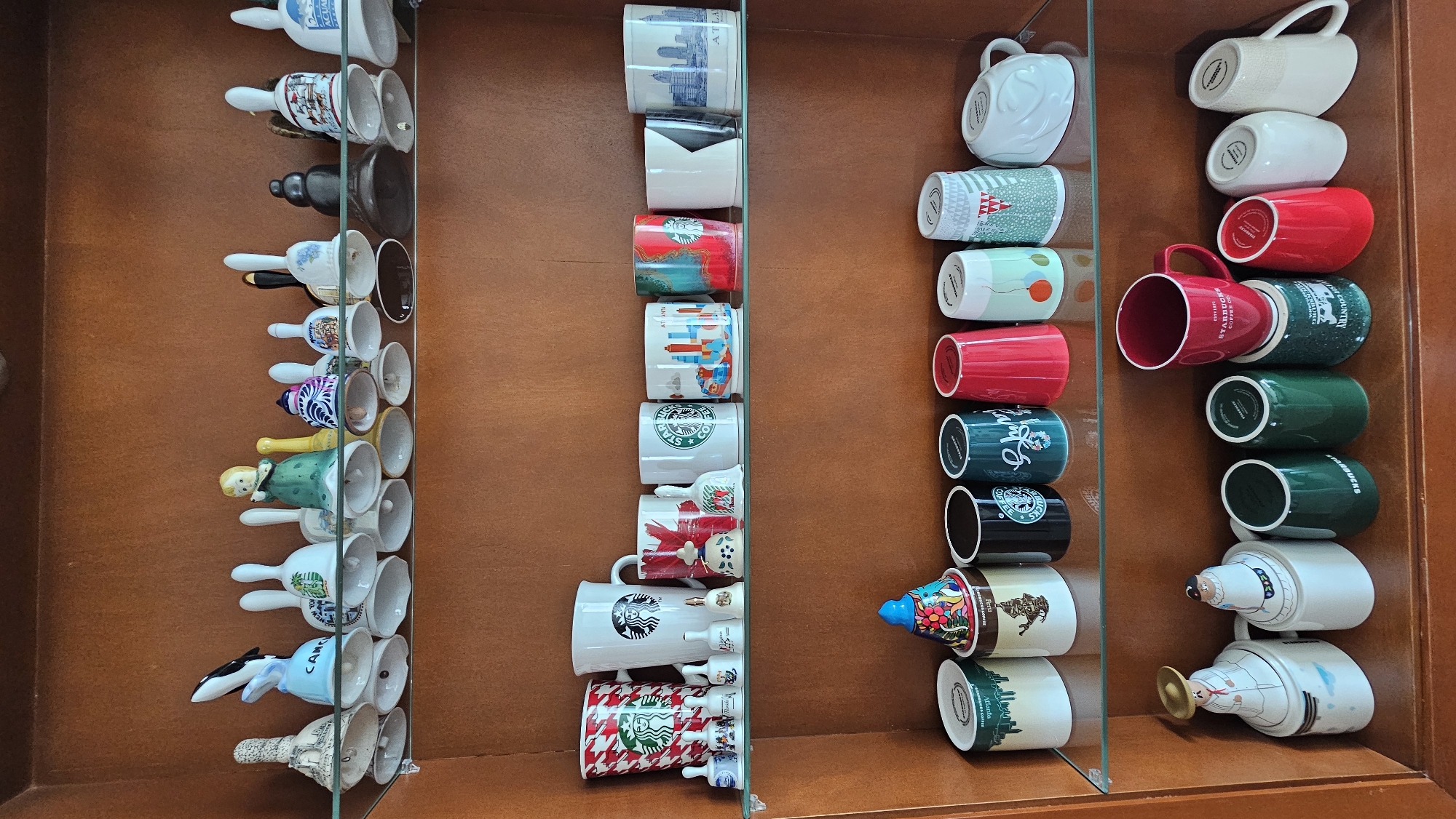 A wooden cabinet displays a collection of coffee mugs and decorative bells on glass shelves. The mugs appear to be Starbucks-branded and depict various cities or themes. The bells are hand-painted with different characters and designs. The scene suggests a curated collection housed in a personal space. No specific details identify the location as San Bernardino Tlaxcalancingo, Mexico.