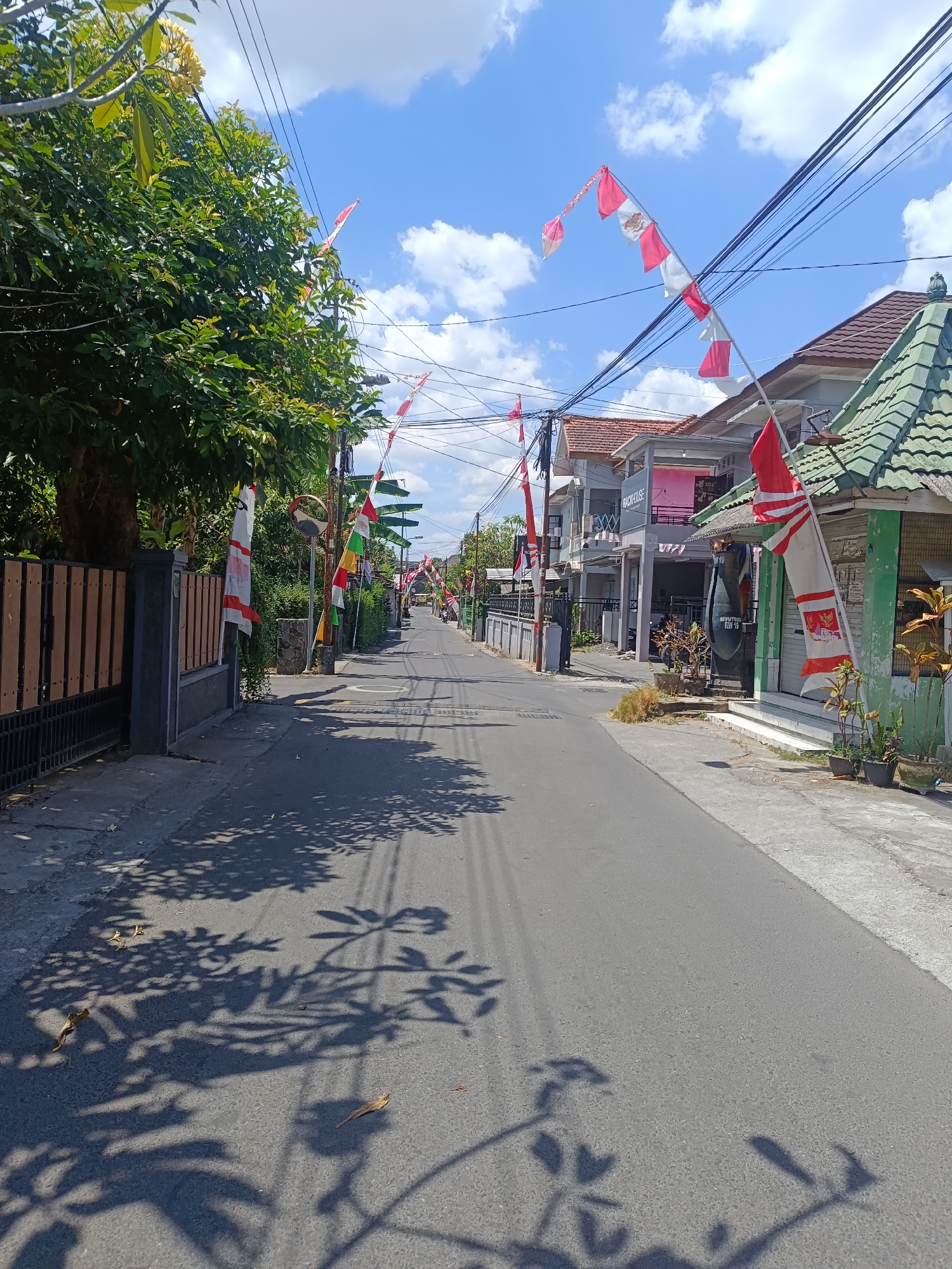The image depicts a street scene in Yogyakarta, Indonesia, likely decorated for a national celebration with numerous red and white flags, characteristic of the Indonesian flag, strung along the street and displayed on buildings. The street is paved and appears to be in a residential or commercial area, with houses and shops lining both sides. Trees with lush green foliage are visible, casting shadows on the road, suggesting it is a sunny day. Overhead, power lines crisscross the sky, a common sight in urban areas of Indonesia. The blue sky is partially covered by white clouds. There are no visible people actively engaged in the scene, although a few birds are present on the road, pecking at something on the asphalt. The overall impression is one of a quiet street, adorned with national symbols, possibly awaiting a celebration.