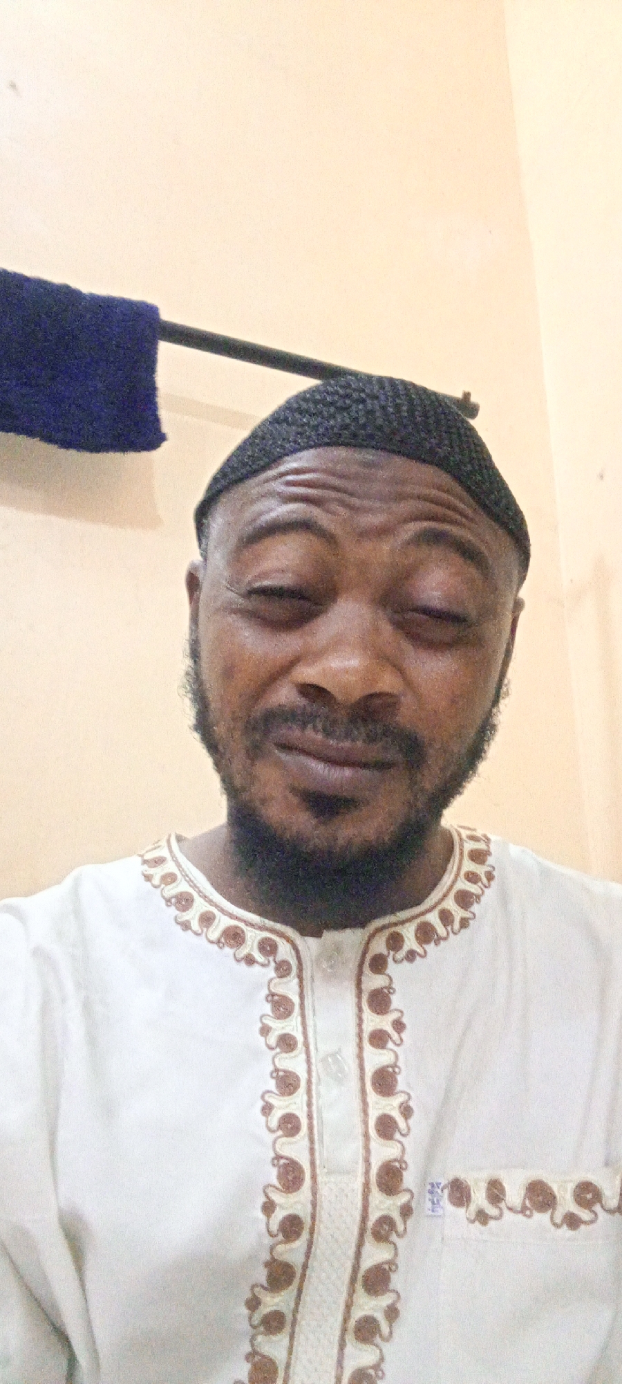 A man is seen in a close-up shot. He has a black crocheted cap on his head, a beard, and is wearing a white shirt with brown embroidery around the collar and down the front. The background is a light-colored wall, with a dark blue cloth draped over a metal bar. The location is Unknown, Unknown.
