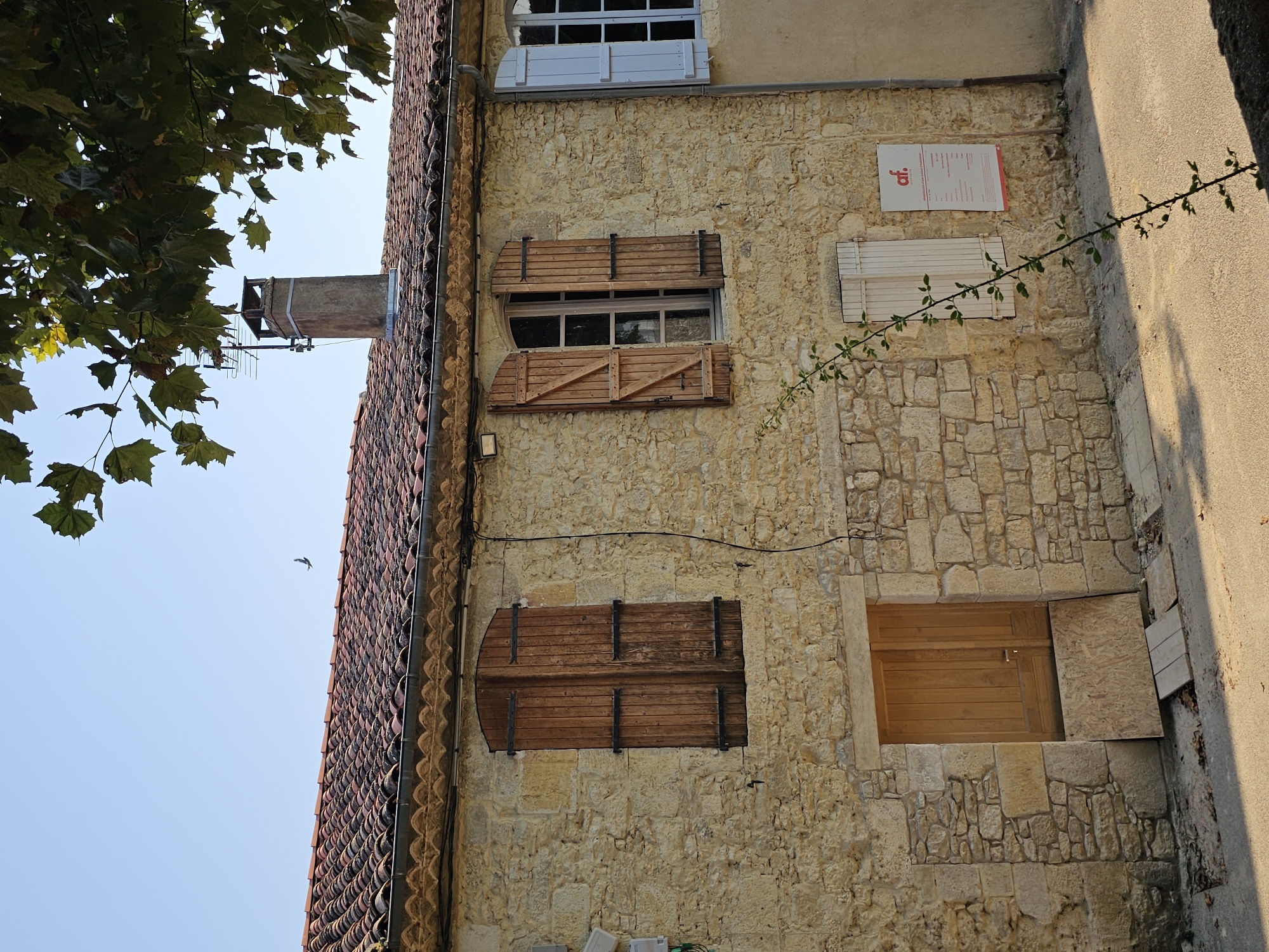 This is an outdoor, eye-level, medium shot of a stone building in the city of Lavardens, France. The building has a red tiled roof, a stone facade, and several wooden shutters. There is a closed wooden door with a piece of plywood leaning against it on the ground floor. Above the door, on the first floor, there are two windows with open wooden shutters. To the right of these windows is another window with closed light blue shutters. A sign with red lettering and white text is mounted on the wall to the right of the open windows. A thin vine with green leaves is growing up the side of the building. In the background, the sky is clear blue, and a small bird is flying. A portion of a tree with green leaves is visible in the upper right corner of the frame. The ground in front of the building is paved.