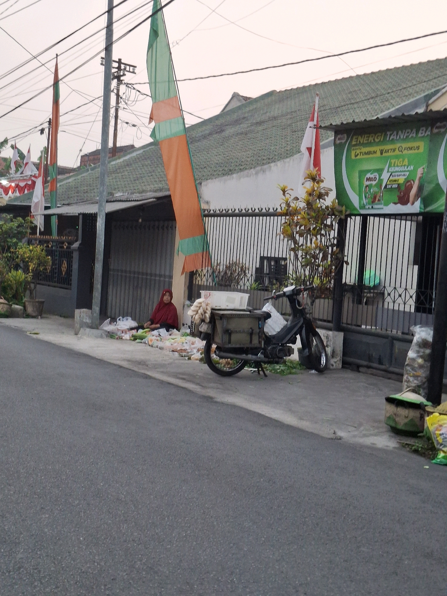 An outdoor, eye-level, medium shot captures a street scene in Lumajang, Indonesia. The foreground is dominated by a gray asphalt road. On the right side of the road, a motorcycle is parked. Behind the motorcycle, there's a black metal fence with vertical bars, and beyond the fence, a building with a green roof and light-colored walls is visible. A large green banner with Indonesian text and an image of a young boy holding a chocolate bar is affixed to the fence. On the left side of the road, a woman wearing a red hijab and maroon clothing is sitting on the ground next to a collection of plastic bags filled with produce, including what appears to be cucumbers and other vegetables. Several flags are displayed on flagpoles in front of the building, with one flag predominantly orange and green, and others appearing to be Indonesian flags. The sky above is a pale, hazy orange, suggesting either early morning or late evening.