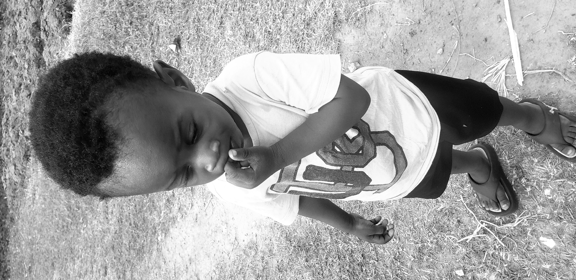 A high-angle, black-and-white medium shot shows a young Black child with short, dark curly hair, wearing a light-colored t-shirt with a large graphic design and dark shorts. The child is standing outdoors on a patch of grass and dirt, with their right hand brought to their mouth, appearing to be sucking on their thumb or finger. The child's head is bowed, and their gaze is directed downwards. They are wearing dark flip-flops. The background consists of dry, patchy grass and dirt, suggesting an outdoor environment in Narayi, Nigeria. The lighting is flat, with no harsh shadows. The scene is static and peaceful.