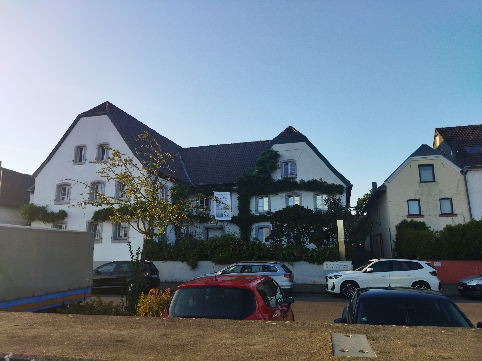 The image shows a street scene in Leverkusen, Germany, featuring a white, multi-story building with a dark tiled roof that appears to be a historical structure, possibly a guesthouse or seminar house given the signage visible. The building is partially covered in green vines. In front of the building, several cars are parked along the street, including a black SUV, a silver station wagon, a red hatchback, and a white SUV. A large, grey truck trailer is also visible on the left side of the frame. The sky is clear and blue, suggesting it is daytime. The foreground is dominated by a low concrete wall, behind which the cars and building are situated.