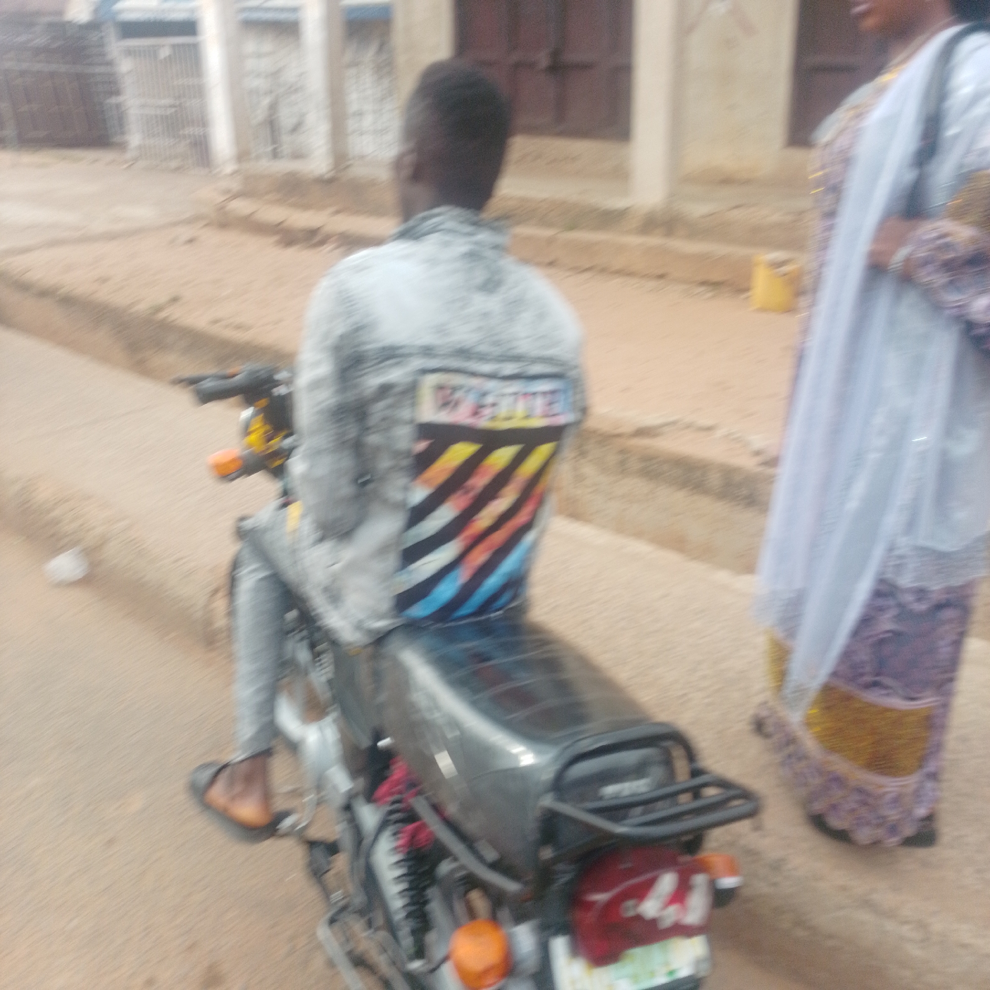 In Garko, Nigeria, a person wearing a grey denim jacket with a colorful striped design on the back and the word "WHITE" is riding a motorcycle. The jacket's design features diagonal stripes in yellow, orange, red, blue, and black. The rider is wearing grey pants and dark open-toed sandals. A second person, partially visible on the right, is wearing a light blue hijab or headscarf and a patterned purple dress with gold accents. The motorcycle is dark grey with a red taillight and an orange turn signal. The ground is a mixture of dirt and sand. In the background, there are buildings with barred windows and doorways, suggesting an urban or semi-urban setting. The overall scene appears to be a street or roadside in Garko.
