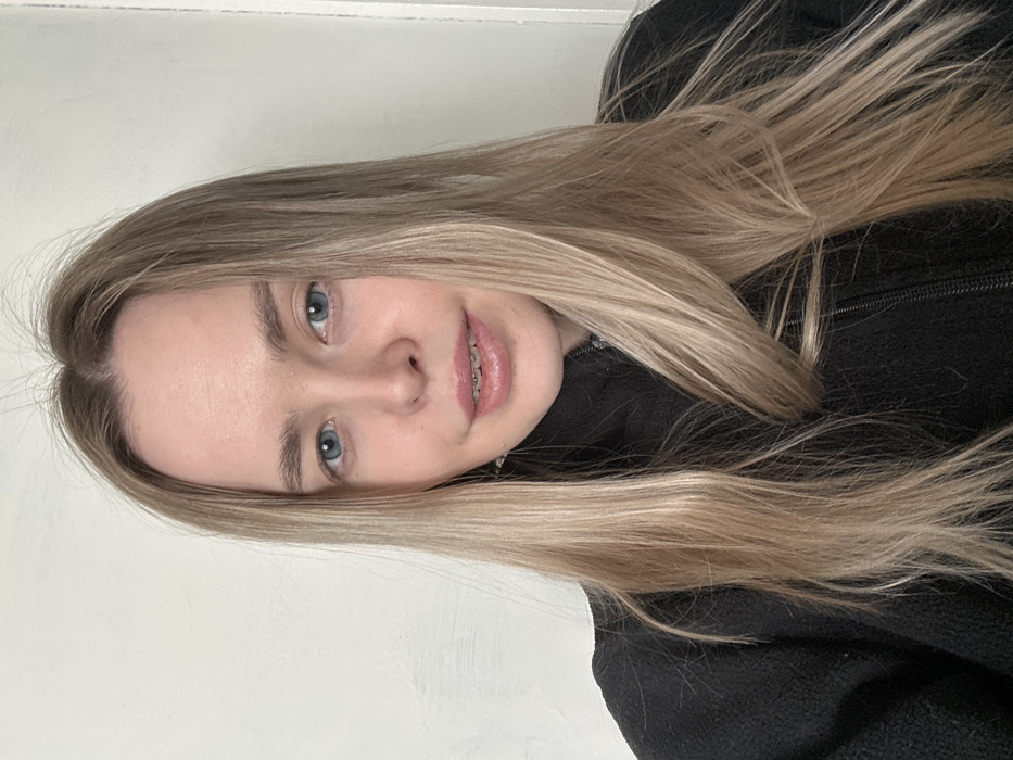 A young woman with light blonde hair and blue eyes is facing the camera. She has braces on her teeth. Her hair is long and straight, falling over her left shoulder. She is wearing a black fleece jacket. The background is a plain white wall. The image is rotated sideways.