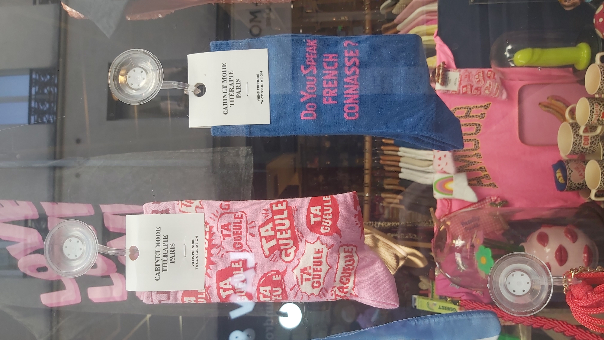 The image is a close-up shot of a retail display window, likely in Paris, France, showcasing a selection of socks and other merchandise. In the foreground, two pairs of socks are prominently displayed. The left pair is pink with repeating red text in speech bubbles that reads "TA GUEULE," a French phrase meaning "shut up." A white tag attached to this pair reads "CABINET MODE THERAPIE PARIS" and "VIENS PRENDRE TA CONSULTATION." The right pair of socks is blue and features pink text that reads "Do You Speak FRENCH CONNASSE?". A similar white tag is attached to this pair, with the same text as the other.
Behind the socks, other items are visible, including a pink t-shirt with the word "AMOUR" printed in a leopard-print font, and a display featuring mugs and other novelty items. The background appears to be the interior of the store, with shelves and racks of clothing. Reflections from outside are visible on the glass of the display window, hinting at an urban environment. The lighting suggests it is daytime.