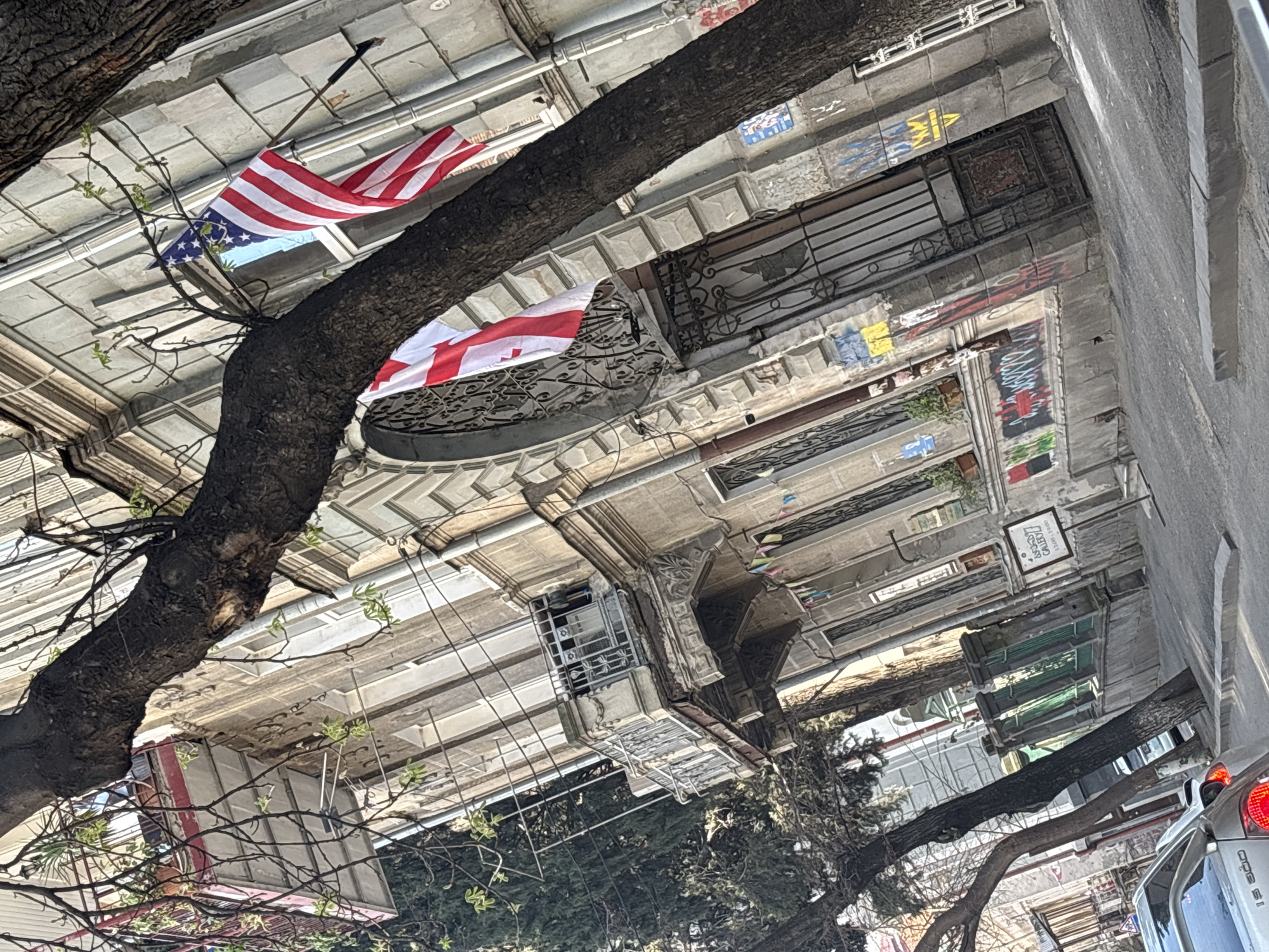 Captured from a ground-level perspective, the image presents an upward view of a building facade in Tbilisi, Georgia. A tree branch with sparse foliage spans the foreground. Above, two flags are visible: an American flag and the flag of Georgia, both mounted on poles attached to the building. The facade shows signs of aging, with visible wear and tear. Several windows and balconies are interspersed along the building's exterior. Graffiti and other markings are present on the walls. Part of a car is visible on the bottom right corner. The overall composition suggests an urban setting, capturing a candid moment of everyday life in Tbilisi.