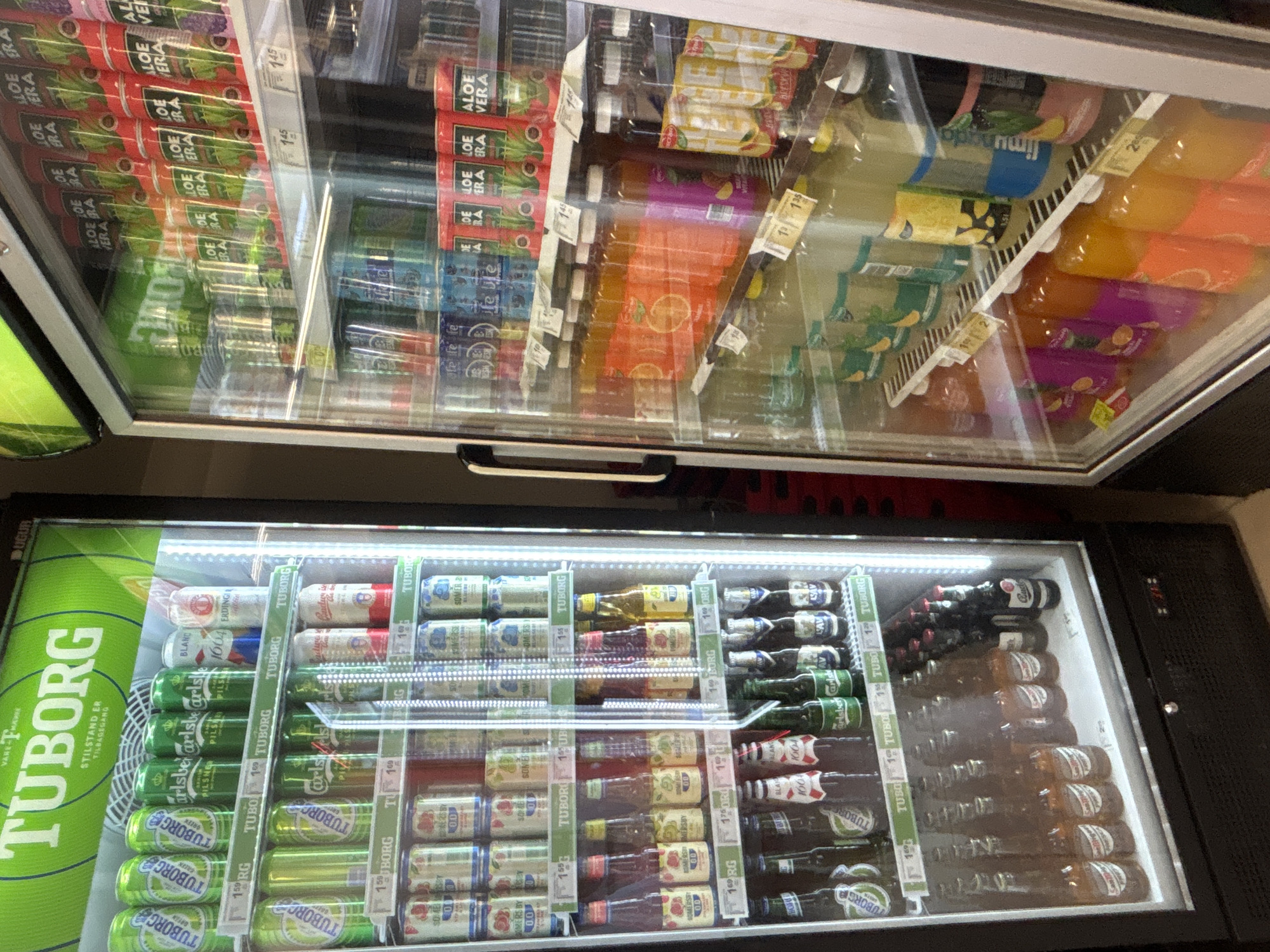 The image shows the interior of two refrigerated display cases stocked with beverages. The top case contains various bottles of juices and sodas with colorful labels, including aloe vera drinks and citrus-flavored beverages. Some bottles are clear, revealing the liquid inside, while others have colored liquid. Price tags are visible on some shelves.
The bottom case is primarily filled with cans and bottles of beer. Prominently displayed is the large green Tuborg logo on the left side of the case. Inside the case, rows of green Tuborg cans are visible, along with other brands like Carlsberg Pilsner and Tuborg Green. There are also bottles of beer and cider, such as Somersby. Some bottles have red and white labels, suggesting specific beer types. Price tags are also present in this section.
The scene appears to be a retail environment, likely a convenience store or supermarket, given the organized display of drinks in refrigerated units. The lighting suggests an indoor setting, possibly during daytime, as there's no indication of natural light. The focus is entirely on the products, with no people or specific environmental details beyond the refrigerator units themselves. The visible text includes brand names like "TUBORG," "Carlsberg," "Somersby," and "ALOE VERA," along with price indicators like "145," "159," and "176."