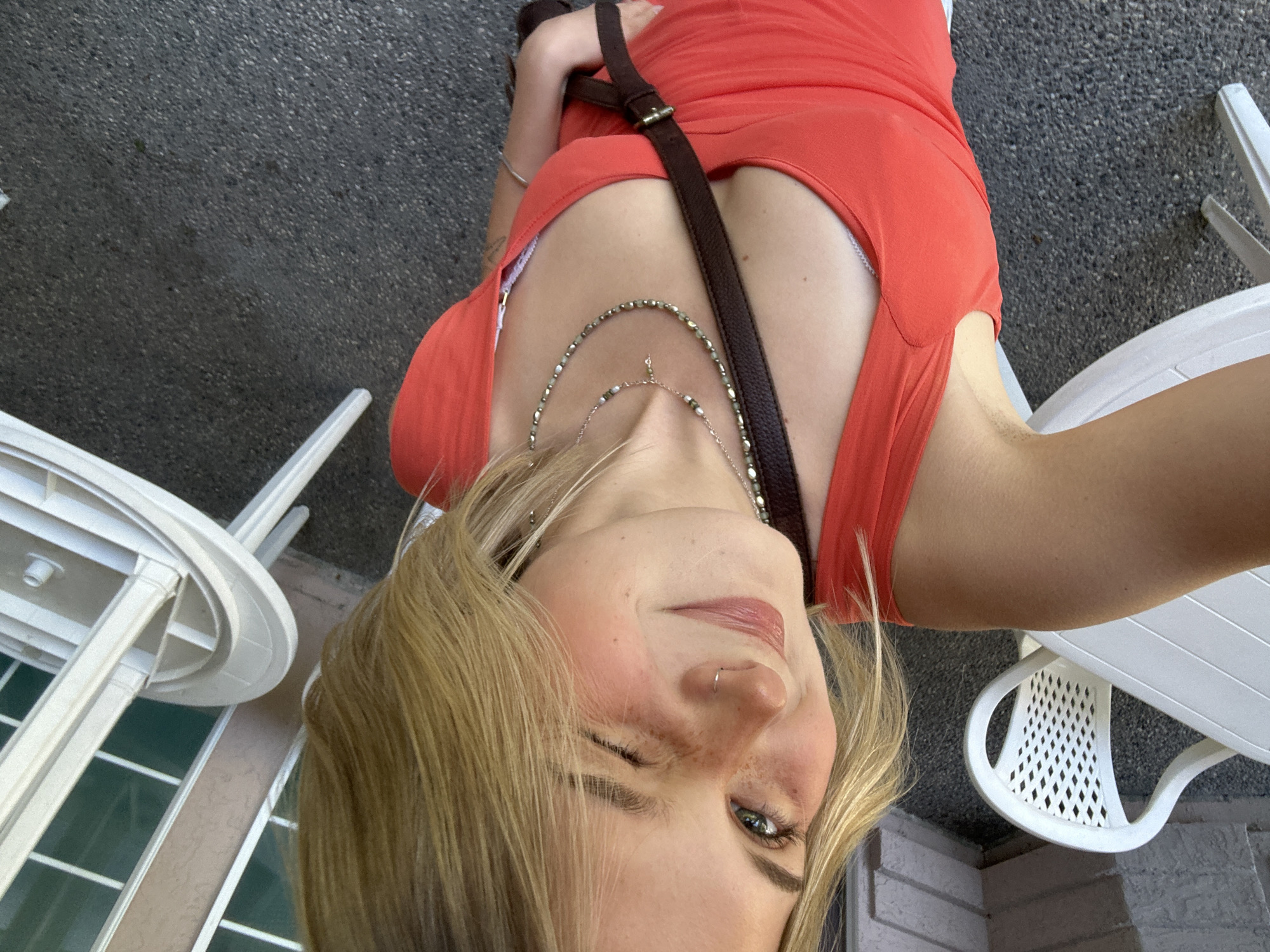 A woman with blonde hair, wearing an orange dress and a brown strap across her chest, is taking a selfie from a low angle. She is winking at the camera. She is wearing jewelry, including a necklace and a nose ring. In the background, there are white plastic chairs and a building with windows. The surface under the chairs appears to be concrete. The location may be Kelowna, Canada, but there are no identifiable landmarks to confirm this.