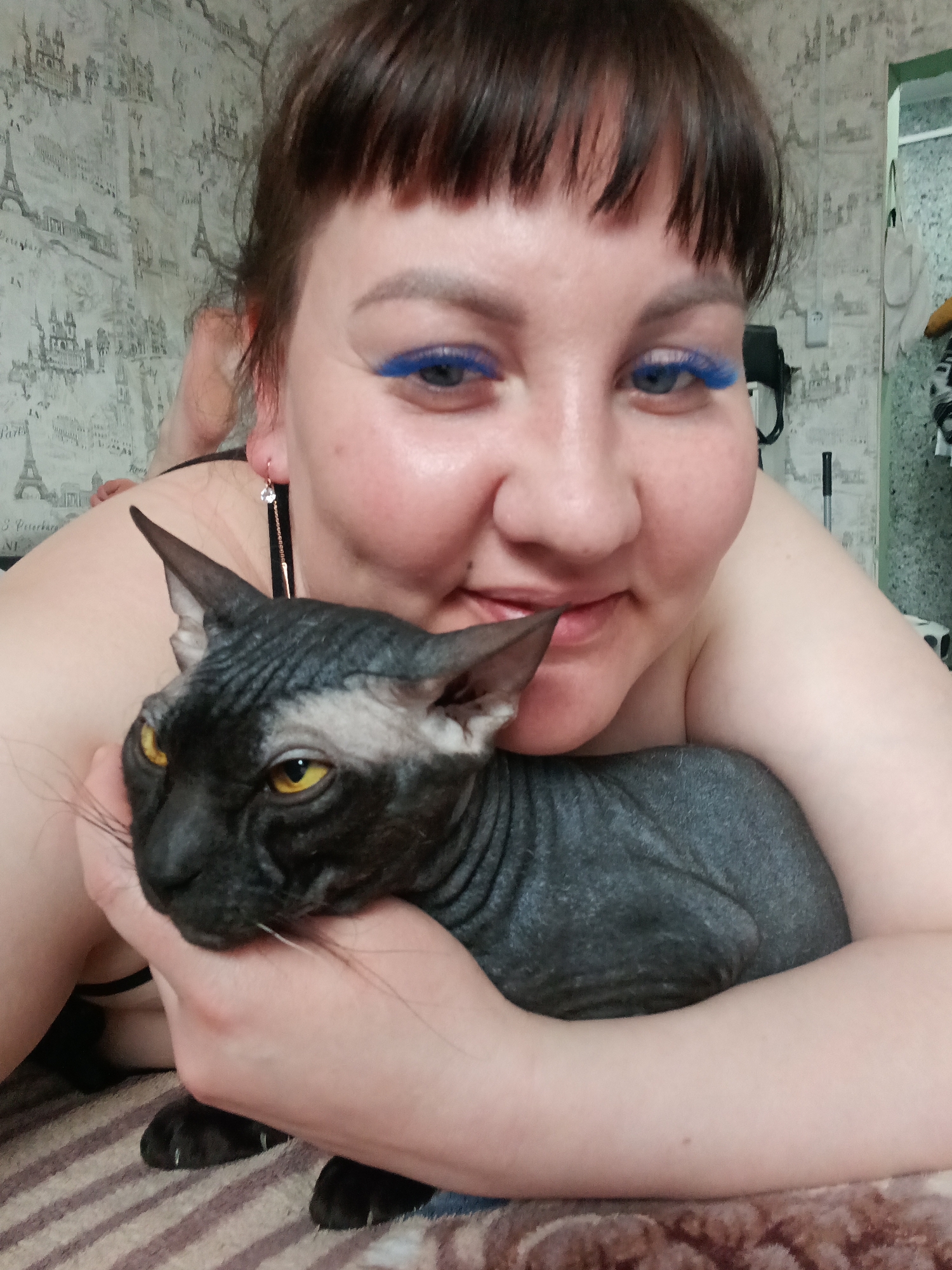 A woman with blue false eyelashes is prone on her stomach, embracing a dark gray Sphynx cat. She has dark brown hair with bangs and is smiling slightly. She has a black top on with a single long crystal earring visible on her left ear. The cat has yellow eyes and is looking to the left. The woman's arm is holding the cat close. The background features a wall with a repeating pattern of the Eiffel Tower and other Parisian landmarks. A partially visible doorway with clothes hanging is to the right. The location in Osa, Russia is unknown.