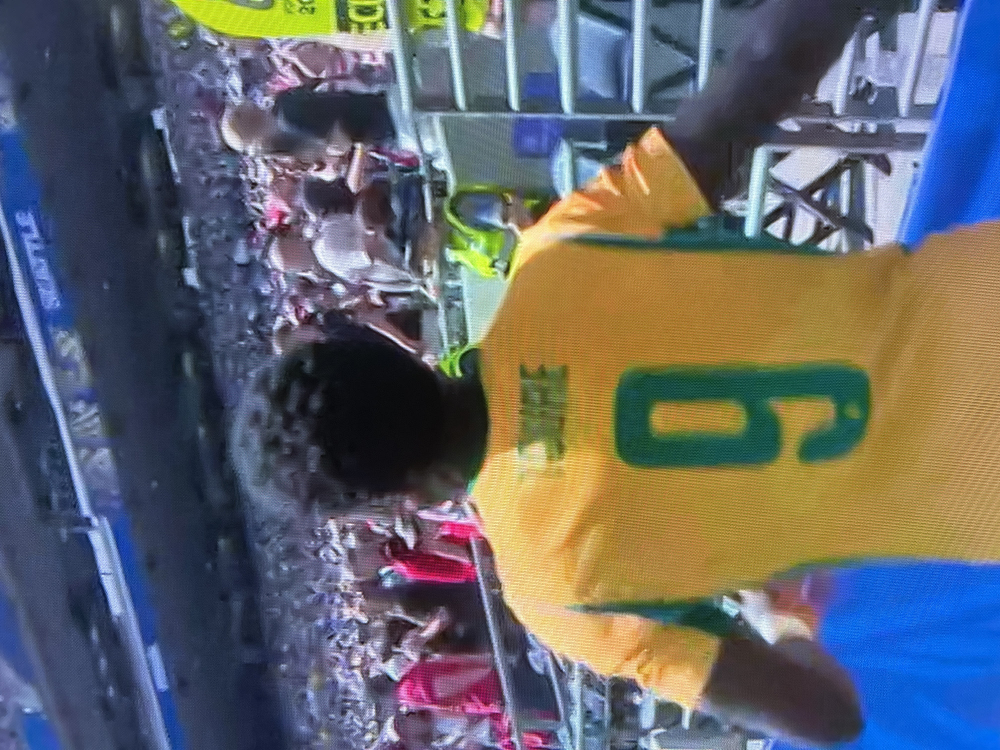 A back view of a person in a yellow jersey with green trim and the number nine is captured in an outdoor setting. The jersey has text above the number which appears to be "SUAREZ". The person is standing in front of a crowd of spectators and what looks like stadium seating. The background includes a blue barrier or pool, and a portion of a structure with shelves and a yellow vest is visible. Above the stadium seating, the word "SEATTLE" is visible on a sign. The lighting suggests it is daytime.