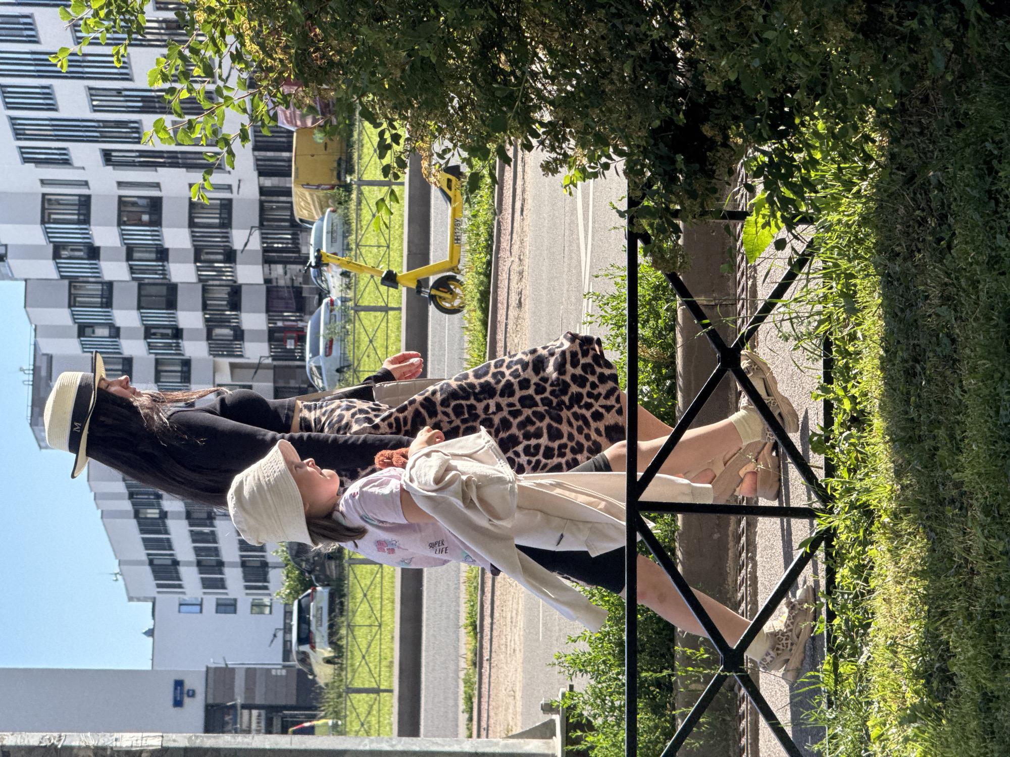 This is a vertical, outdoor, medium-shot photograph taken during the day, under sunny weather. The scene depicts two people, an adult and a child, walking alongside a road. The adult, in the foreground, wears a wide-brimmed straw hat with a dark band, and a black top with a leopard-print skirt. The child, positioned in front of the adult, wears a light-colored sun hat and a pink t-shirt with some graphic text. Both individuals appear to be casually dressed, suggesting a relaxed outing.
The background reveals a modern apartment building with a distinctive checkered facade, indicative of contemporary architecture. Beside the road, a yellow electric scooter is visible, suggesting a mode of shared transportation common in urban environments. Further in the background, cars and more buildings can be seen, reinforcing the urban setting. A black metal fence runs along the edge of the grassy area where the people are walking, separating them from the road.
The lighting suggests it is daytime, likely late morning or afternoon, with distinct shadows cast by the foliage and the fence. The overall mood appears peaceful and summery. The text "SUPER LIFE" is visible on the child's t-shirt. Based on the context provided, the location is Saint Petersburg, Russia.