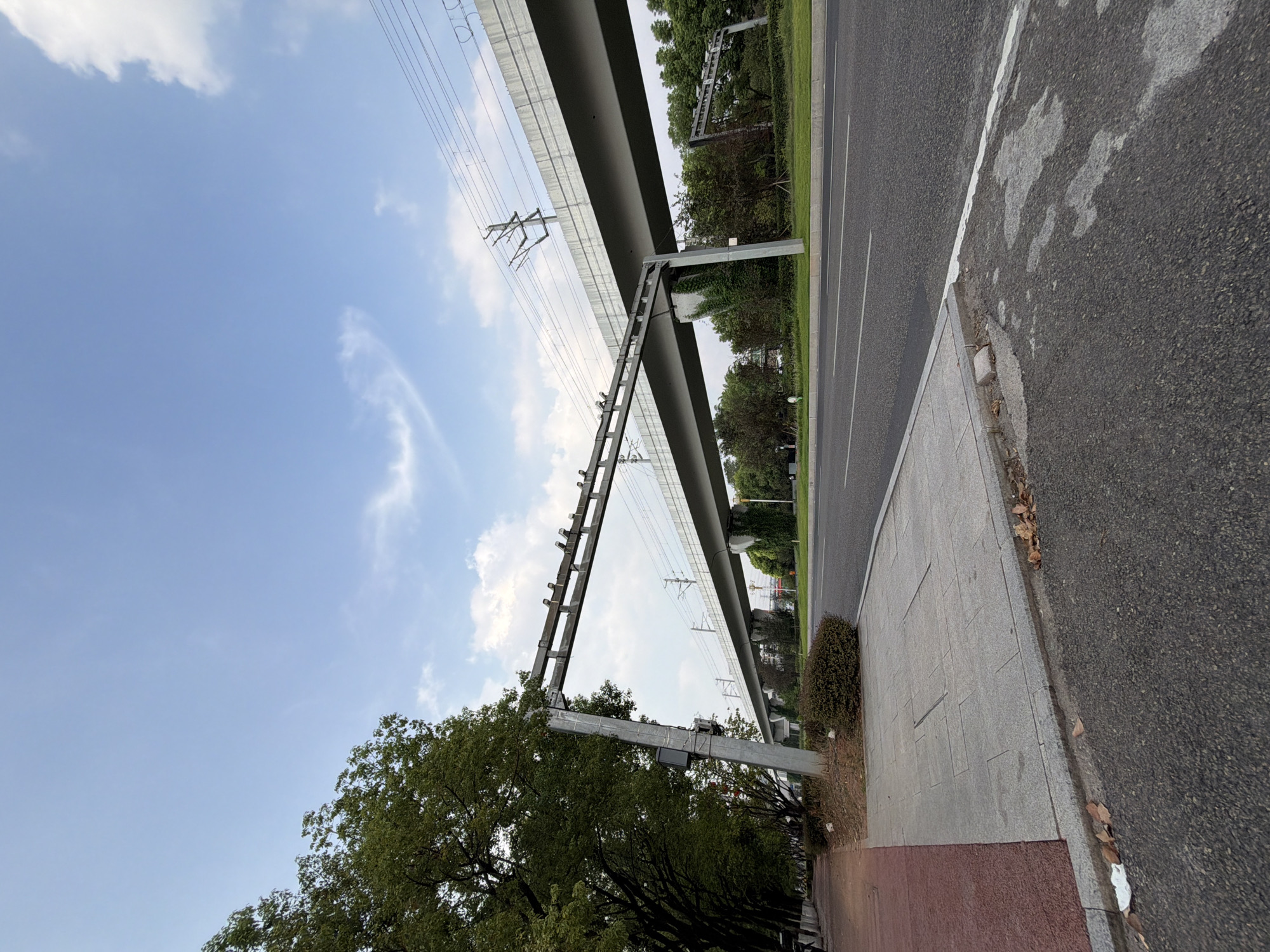 In Jiangjin, China, a wide asphalt road runs parallel to a sidewalk, lined with trees. A raised monorail track structure, supported by concrete pillars, stretches across the scene, with overhead power lines visible. The sky above is a clear blue with scattered clouds. The scene appears to be a public thoroughfare, possibly near a transportation hub. No people are present.