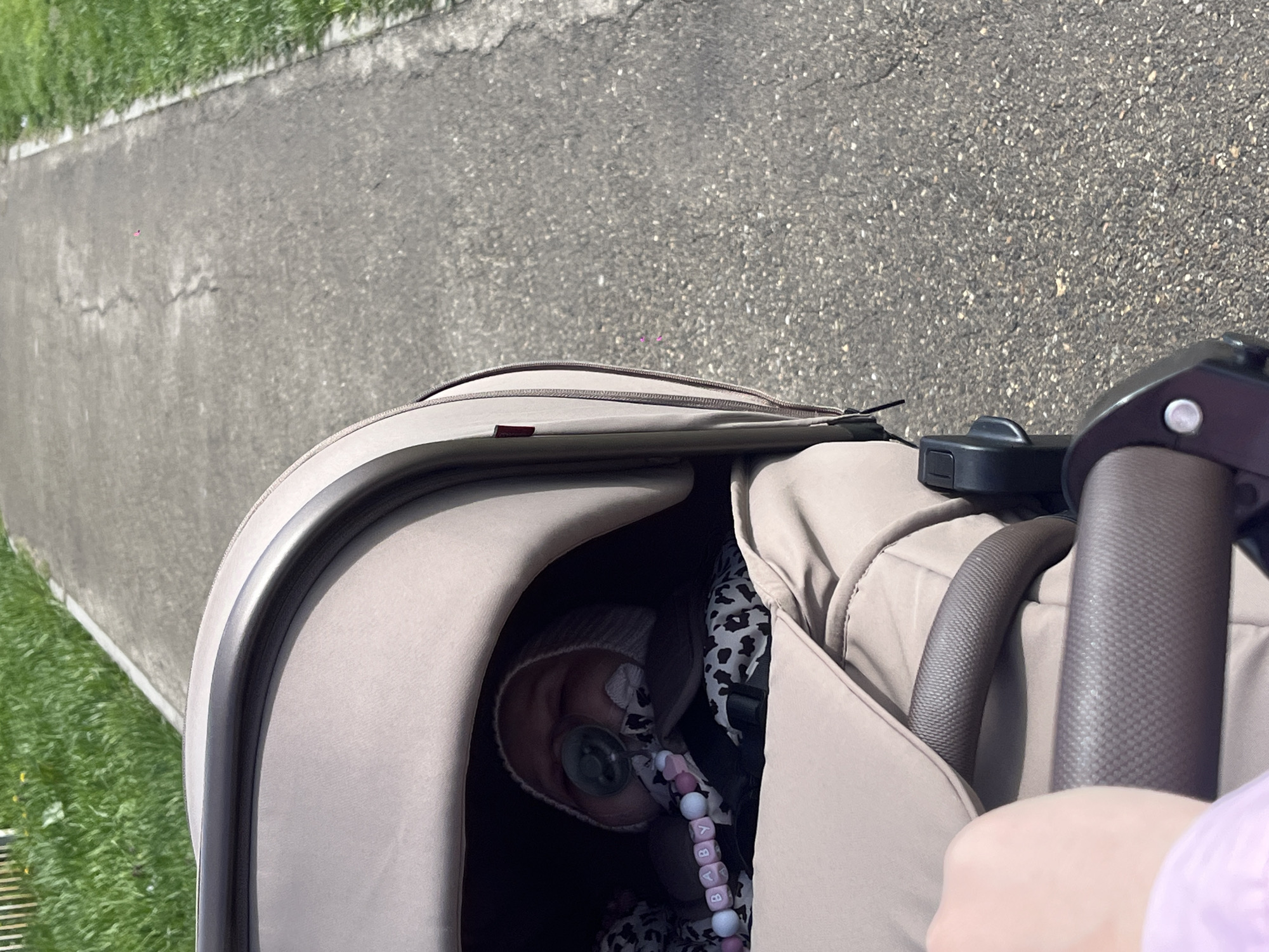 A top-down, eye-level shot shows a baby in a stroller on a sidewalk. The baby, wearing a beanie, has a pacifier in its mouth. A beaded pacifier clip spelling out "BABY" is visible. The stroller is beige and has a dark interior. A sidewalk is adjacent to a grassy area. The exact location in the city of Novopokrovskaya, Russia, cannot be determined from the image.