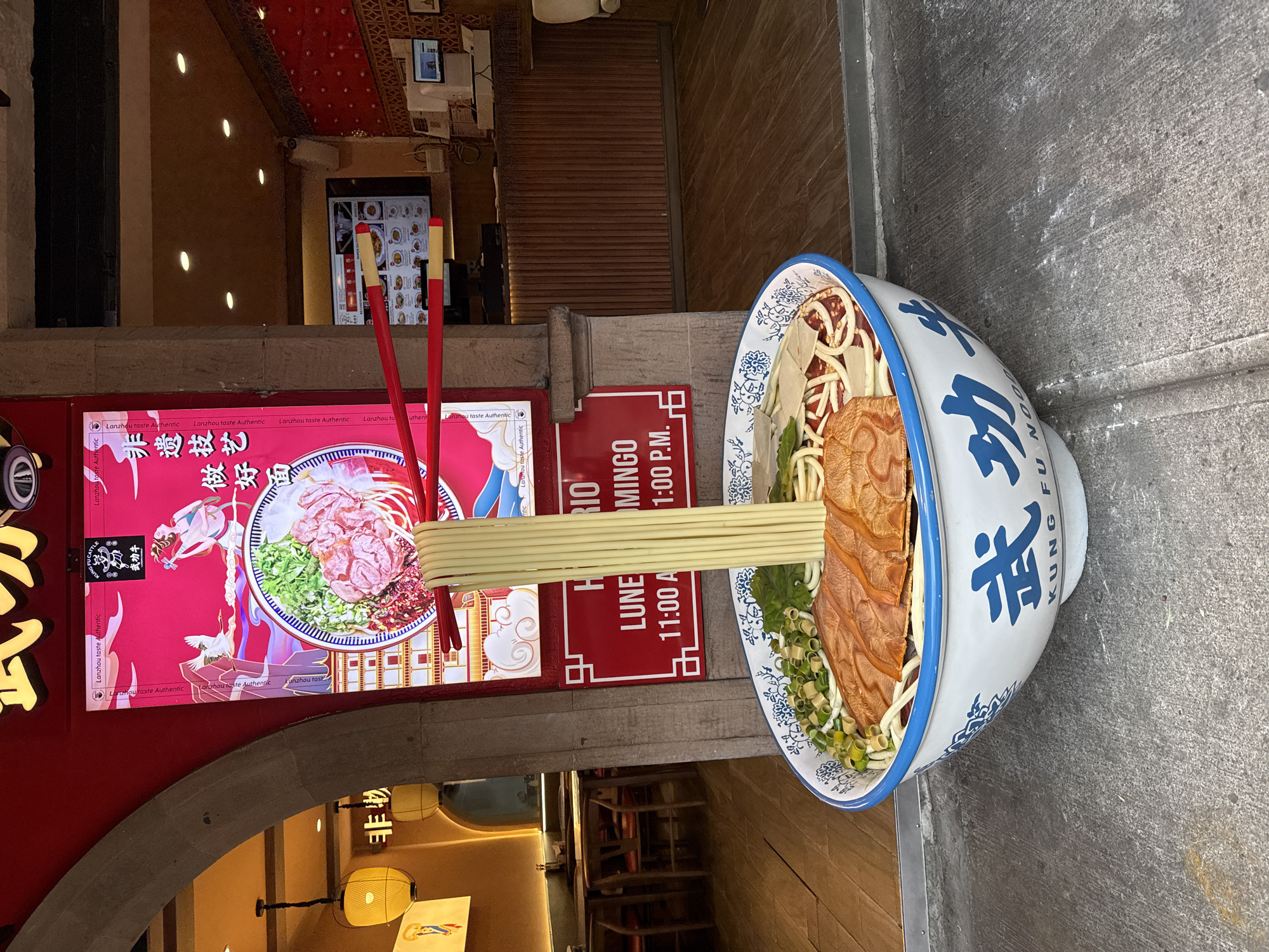 Here is a detailed description of the image:
**Overall Impression:** The image shows a restaurant exterior with eye-catching food-related decorations, including signs and a large bowl of noodles.
**Location:** The location is in Mexico City, Mexico, implied by the signage and potentially by architectural styles.
**Subjects/Objects:**
* **Restaurant Exterior:** The exterior is decorated with red signs featuring Chinese characters and images of food. The signs highlight the authenticity of the cuisine, referring to "Lanzhou taste Authentic" and mention 武功牛 (Kung Fu Cattle). A vertical red sign displays the restaurant's opening hours for "LUNE" (Monday) and "MINGO" (Sunday).
* **Giant Bowl of Noodles:** A large, three-dimensional bowl filled with noodle ingredients is placed prominently. The bowl has blue Chinese characters written on its white exterior.
* **Chopsticks:** Oversized red chopsticks are visually integrated with the signs.
* **Archway:** The restaurant entrance is accessed through a stone archway.
* **Interior View:** Through the archway, the restaurant's interior is visible, with seating, lighting fixtures, and stairs leading to another level.
* **Menu Screens:** Electronic menu screens are positioned inside the restaurant near the front.
* **Sidewalk:** The restaurant's entrance is placed on a sidewalk with concrete.
**Environment:**
* The location appears to be in a mixed-use area with both commercial and residential buildings.
**Overall:** The scene captures a restaurant that aims to present an authentic experience of Chinese cuisine. The decorations are designed to attract attention and communicate the restaurant's specialization in Lanzhou-style noodles.