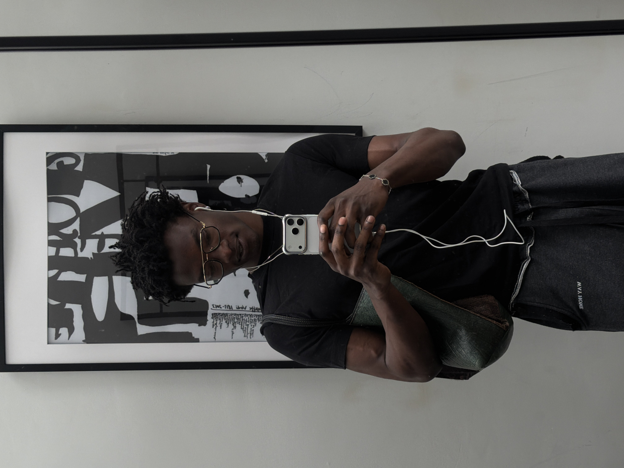 A Black man with dreadlocks and gold-rimmed glasses is taking a selfie in front of a framed abstract artwork. He is wearing a black t-shirt, grey sweatpants, and white earbuds. A dark green purse is slung over his left shoulder. A silver bracelet with black clover-shaped charms adorns his right wrist. The background is a light-colored wall with a framed black and white print. The scene appears to be indoors, possibly in an art gallery or a home.