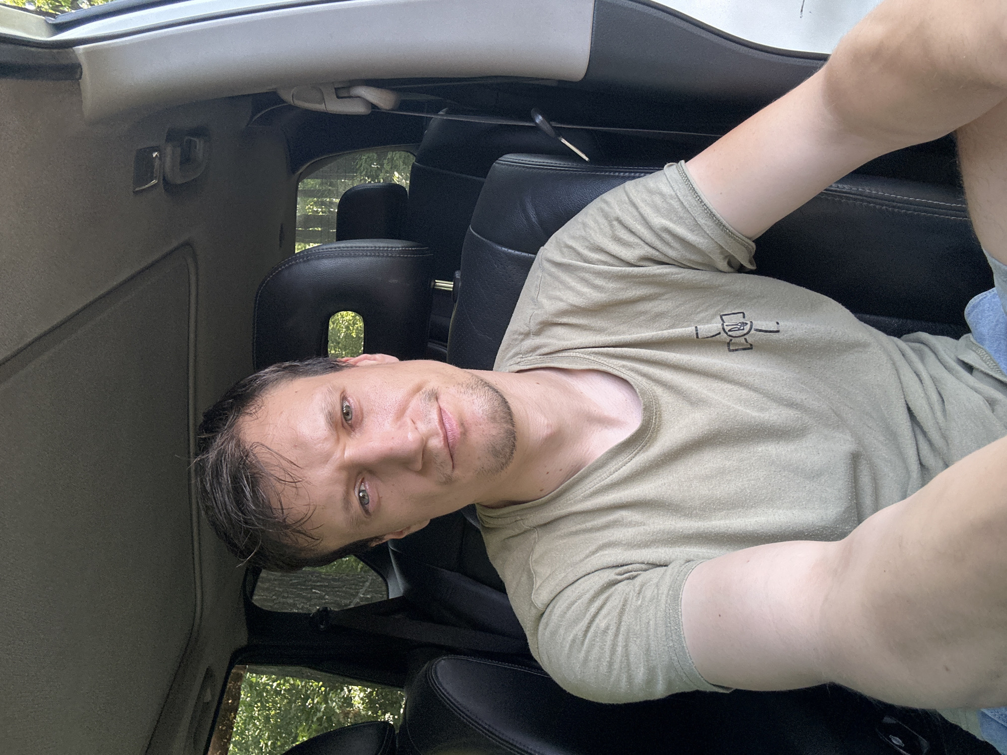 A man is sitting in the driver's seat of a car, taking a selfie. He is wearing a light green t-shirt and blue shorts. The car appears to be parked on a dirt road, with trees and foliage visible through the windows. The interior of the car is dark, with black leather seats and a sunroof. The man's face is visible, and he has short brown hair and green eyes. He is looking directly at the camera with a neutral expression. The scene is illuminated by natural light.