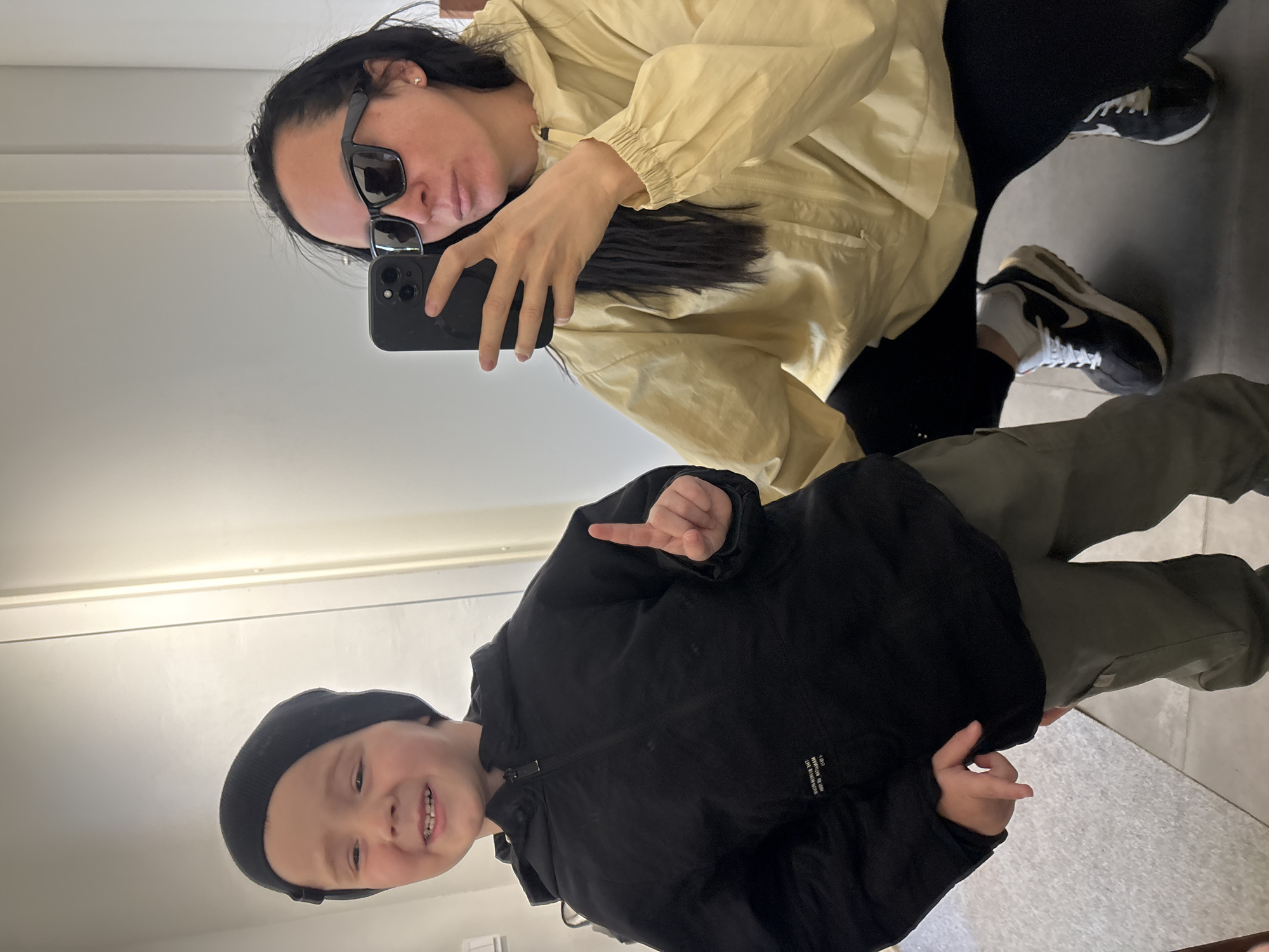 A woman with sunglasses is taking a selfie with a boy wearing a black beanie and jacket. The woman is wearing a light yellow jacket, black pants, and black and white sneakers. The boy is pointing his finger towards the woman. The location appears to be indoors with a light-colored wall and floor. There is no information to suggest this photo was taken in the city of Chaikovsky, Russia.