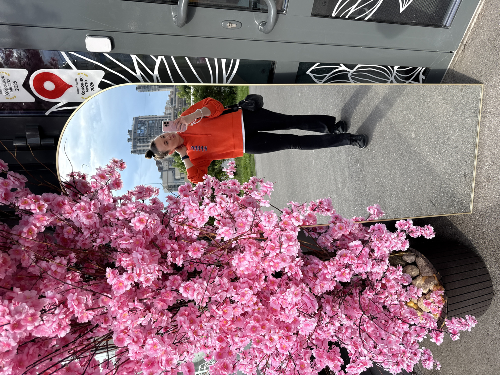 An arched mirror, approximately 6 feet tall, is positioned outdoors on asphalt, reflecting a young woman in an orange hoodie and black pants. She is taking a selfie with a pink smartphone, her hair pulled up in a bun, and a black shoulder bag. To the left of the mirror, a large, decorative tree with abundant pink blossoms is in a dark planter. The background shows buildings and a cloudy sky. On the right side of the mirror, a grey door with decorative white patterns and door handles is visible, along with two stickers on the door. One sticker features a red map pin and the text "Яндекс Карты Хорошее место 2025" (Yandex Maps Good Place 2025), and another sticker below it reads "Яндекс Карты Хорошее место 2026" (Yandex Maps Good Place 2026). This suggests the location is likely a business or establishment in Saint Petersburg, Russia, that has been recognized by Yandex Maps.
