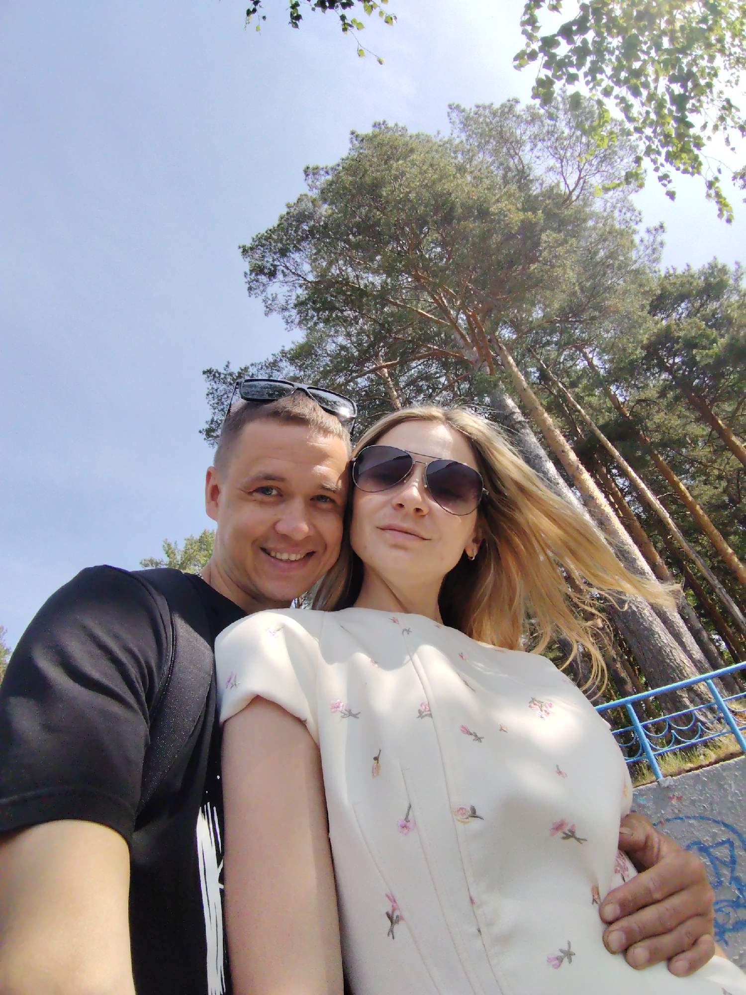 A selfie-style photograph captures a couple standing outdoors with tall trees in the background under a clear sky. The man on the left wears a black t-shirt and has sunglasses perched on his head. He is smiling at the camera. The woman is wearing sunglasses and a light-colored dress with a floral print. Her blonde hair is blown to one side. The man has his arm around the woman. The location is outdoors in the city of Chaikovsky, Russia.