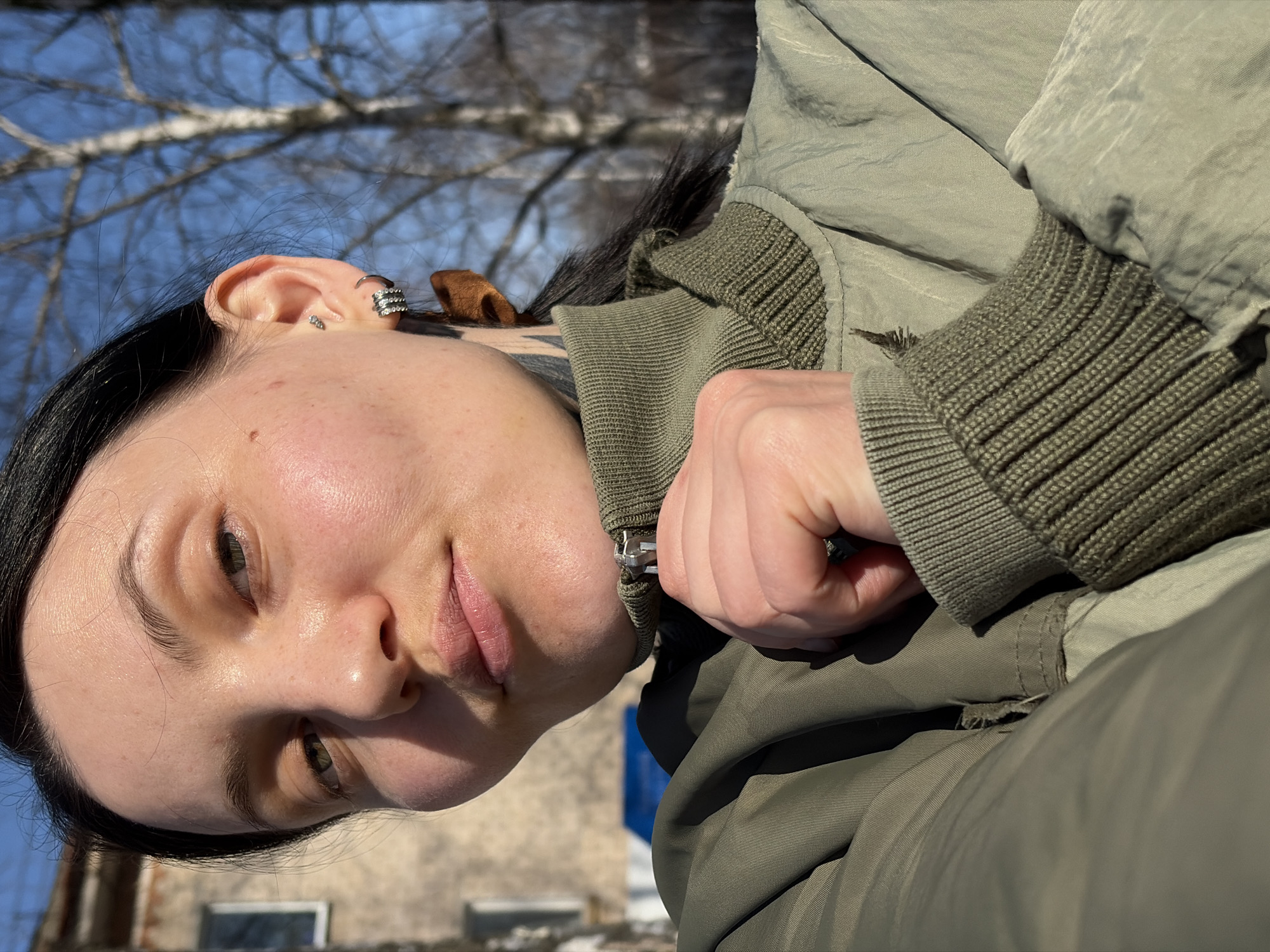 A young woman with dark hair is the main subject of this outdoor photograph. She is wearing a military green jacket with ribbed cuffs and is seen from a slightly downward angle. The background is blurred, but bare tree branches against a light blue sky are visible in the upper left corner. The woman's face is turned to the side, and she has multiple earrings in her left ear. Parts of a building can be seen behind the woman, suggesting a location in a built-up area, possibly Chaikovsky, Russia. The overall lighting appears to be natural, indicating a daytime shot.