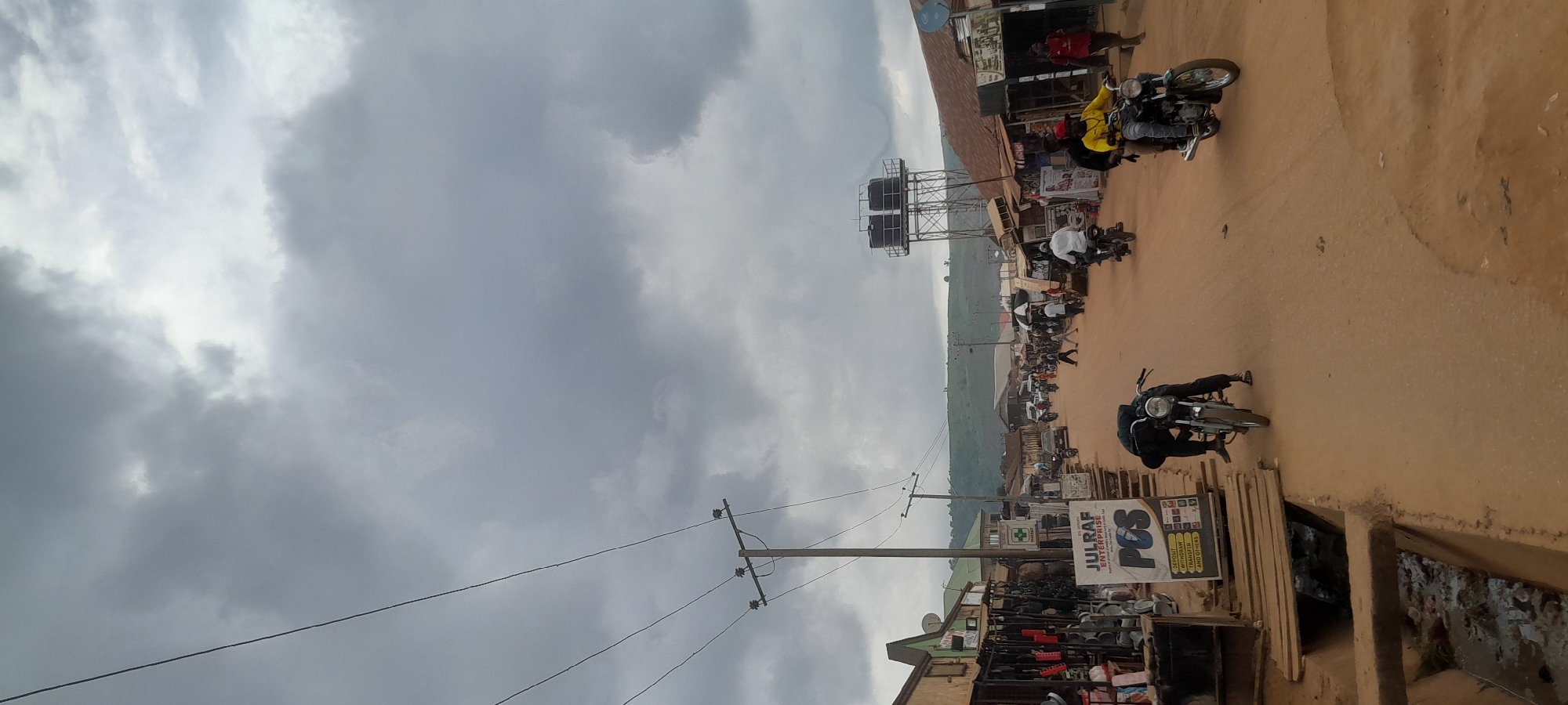 The image depicts a street scene in Abuja, Nigeria, under a heavily overcast sky. The street is unpaved and dusty, lined with shops and stalls on both sides. In the foreground, a person in dark clothing is bent over a motorcycle, appearing to adjust it. Further down the street, several other motorcycles with riders are visible, navigating the road. Some pedestrians can be seen walking along the street and in front of the shops. On the left, a shop displays various goods, and a sign for "JULFRAF ENTERPRISE" with "POS" is prominently displayed. On the right side of the street, more shops are visible, and a water tower stands in the background. The overall atmosphere suggests a bustling local market or commercial area in a developing urban setting. The landscape in the distance shows rolling hills.