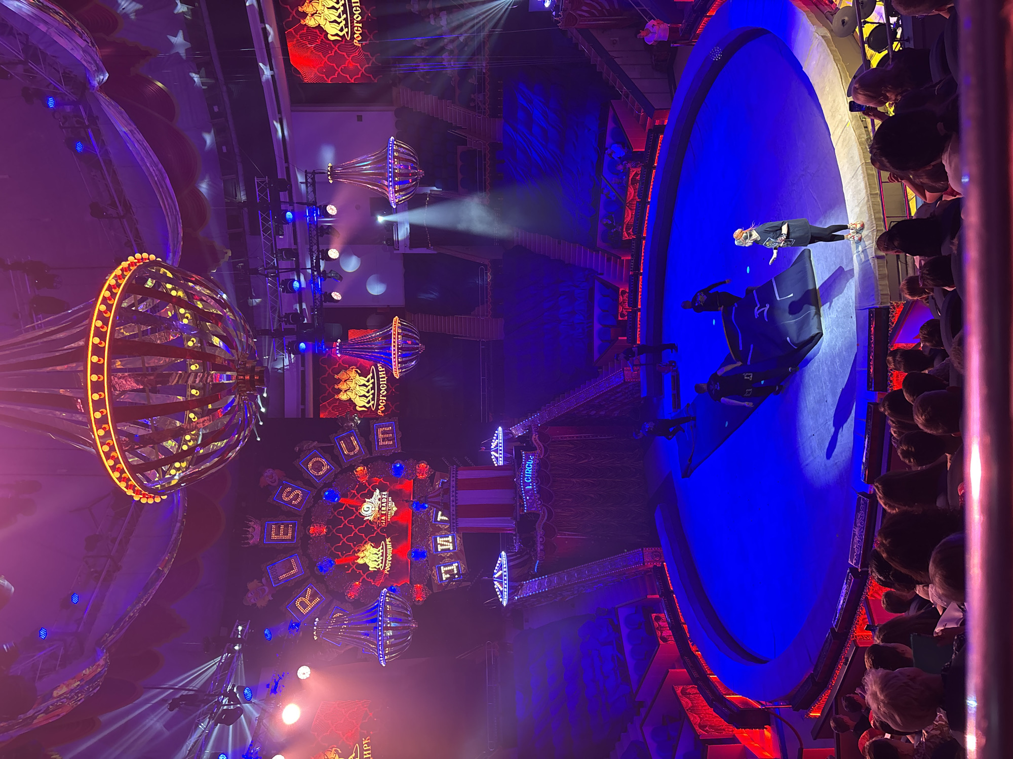 The image shows the interior of the "Royal Circus" in Sochi, Russia. The circus tent is brightly lit with a mix of red and blue lights, creating a vibrant and festive atmosphere. In the center of the tent is a circular stage, with a blue mat laid out. A woman in a black t-shirt and leggings is standing on the stage, facing the audience. She appears to be addressing the audience or giving instructions. Several other individuals, possibly circus performers or crew members, are also present on or near the stage. The audience is seated in tiered seating that surrounds the stage. There are also large chandeliers hanging from the ceiling, adding to the grandeur of the circus. The overall scene depicts a lively and energetic circus performance in progress.