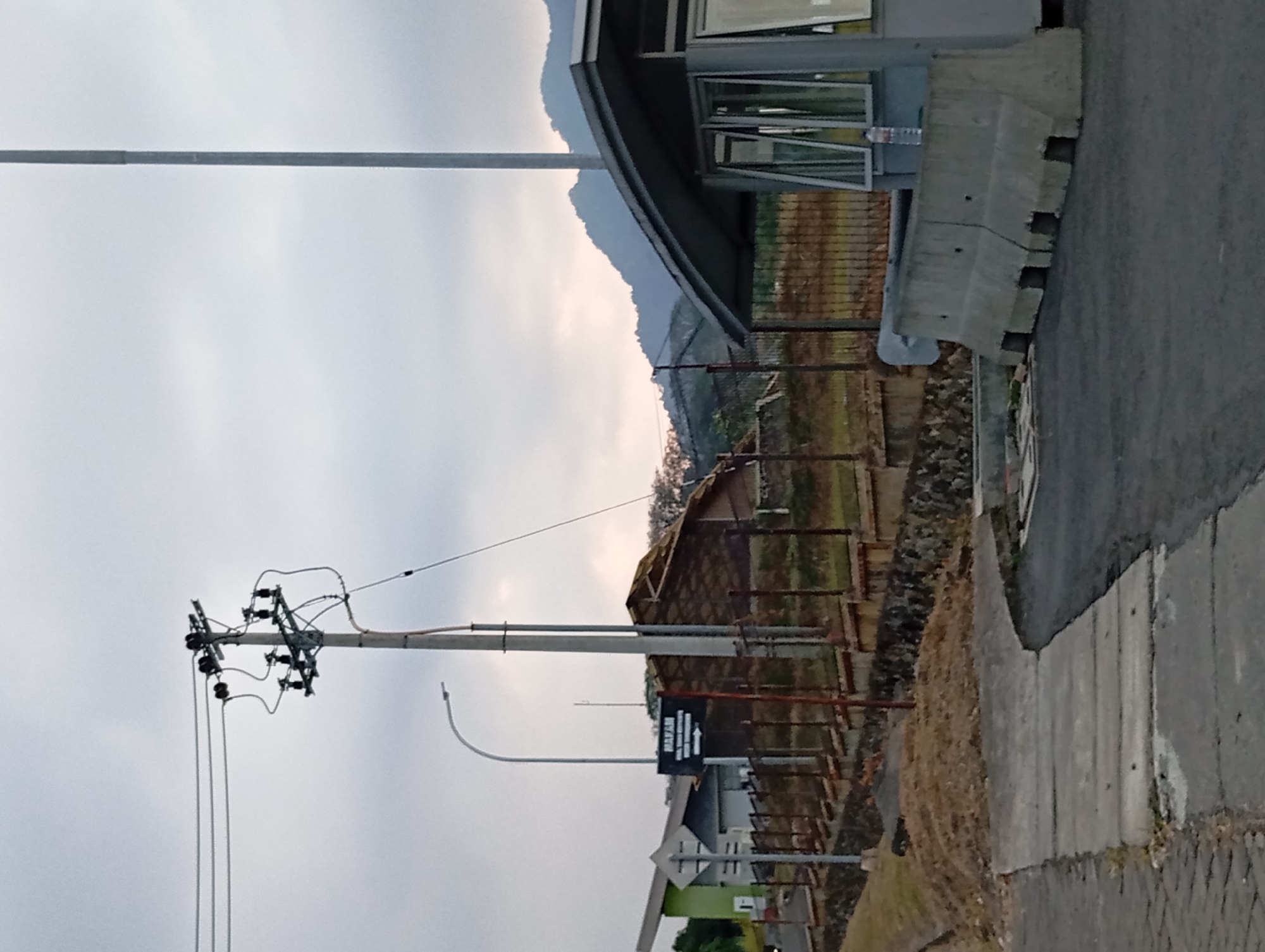 The image shows an outdoor scene in Sumber Suko, Indonesia, likely near a cemetery or memorial. In the foreground, a paved road curves to the right, with a concrete sidewalk or pathway to the left. Two large, precast concrete barriers are positioned on the road, one immediately in front of a gray guard booth. The guard booth has windows and a sloped roof. Behind the barriers and booth, a chain-link fence runs along a raised area covered with dry grass and some rocks. Beyond the fence, several buildings are visible. One building on the left has a green section and a white section. Another structure further back has a roof made of what appears to be corrugated material. A dark sign with white lettering reading "MAKAM" (which means "grave" or "tomb" in Indonesian) and an arrow pointing to the right is mounted on a pole near a streetlamp. In the background, hills or mountains are visible under a cloudy sky. A utility pole with electrical wires and insulators stands prominently on the left side of the image. The overall lighting suggests it is late afternoon or early morning with overcast conditions. There are no people visible in the image.