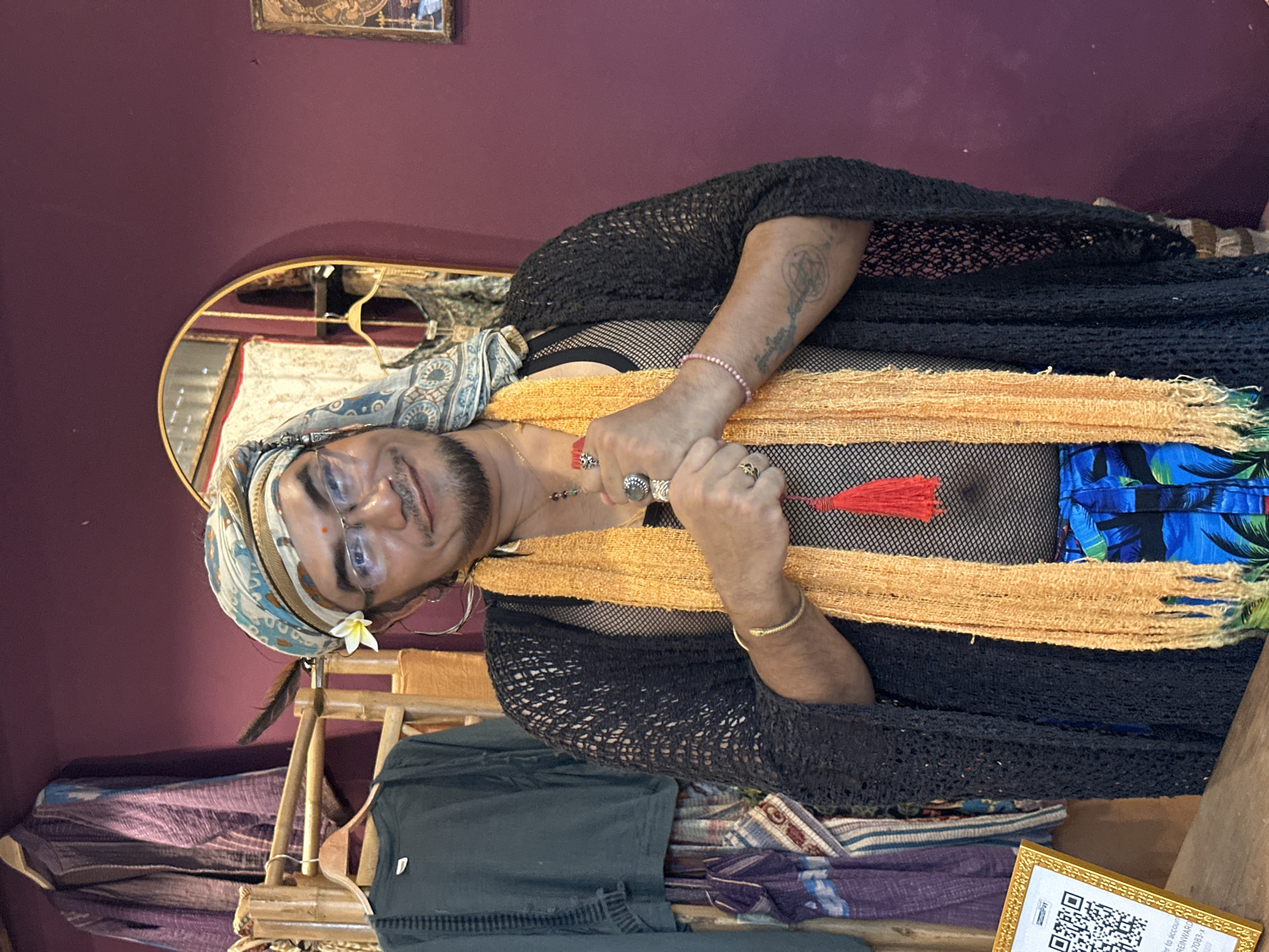 The image shows a person indoors in ตำบลเกาะพะงัน, Thailand. The person is wearing a patterned headscarf with a flower and feather tucked into it, glasses, hoop earrings, a black fishnet top, and a black crocheted shawl. They have an orange scarf draped around their neck and are wearing rings and bracelets. Their arms are crossed in front of them. Behind them is a maroon wall with a picture frame and a mirror reflecting the scene. Below them is a bamboo structure with folded textiles and clothing. A framed paper with a QR code is also visible.