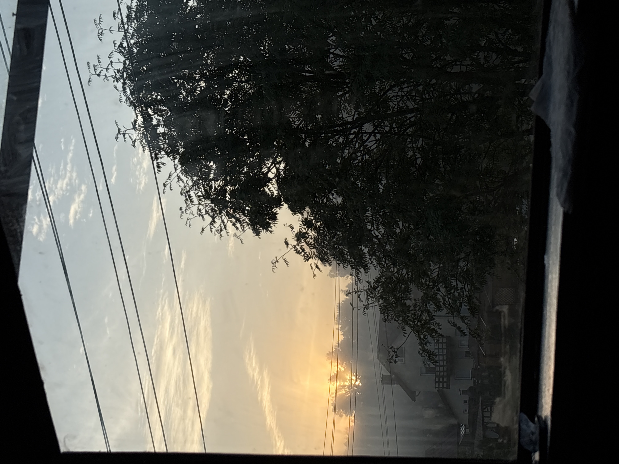 The image depicts a view through a window of a rural landscape at dawn. The sun is rising, casting a warm orange glow on the sky and illuminating a patch of mist. Power lines crisscross the sky, and a large, dark tree dominates the right side of the frame. In the distance, a house with a chimney and a balcony is visible, along with several small outbuildings. The overall impression is one of a peaceful morning in a village setting. The location is identified as Jāņupe, Latvia.