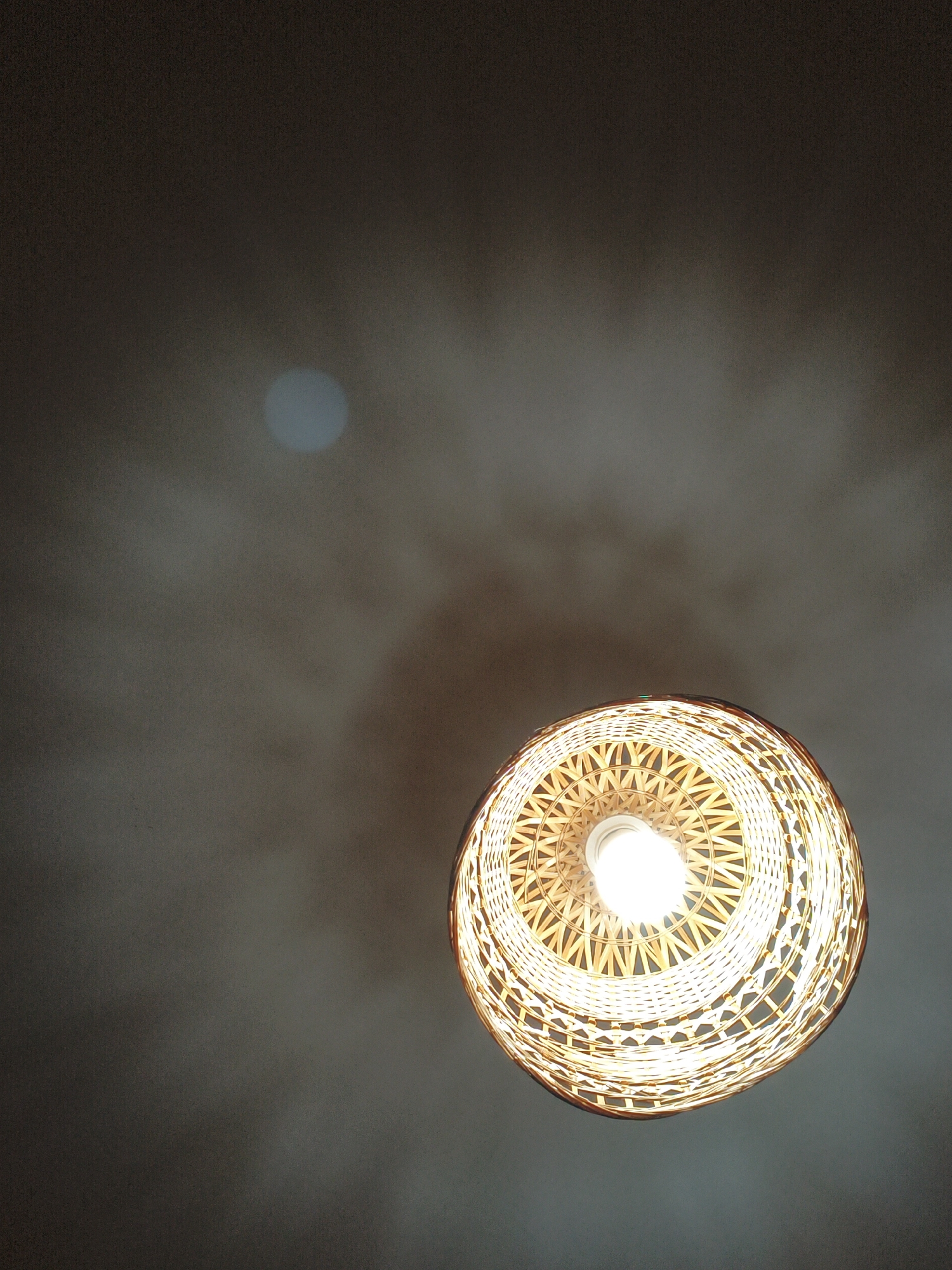 An upward view captures a woven bamboo lampshade with a lit bulb inside, casting a warm glow. The lampshade is suspended against a dark, textured ceiling. To the left of the lampshade, a circular reflection of light is visible on the ceiling. The scene appears to be an interior setting in Lyubertsy, Russia, possibly a room in a residential or commercial building. No people or explicit actions are depicted.