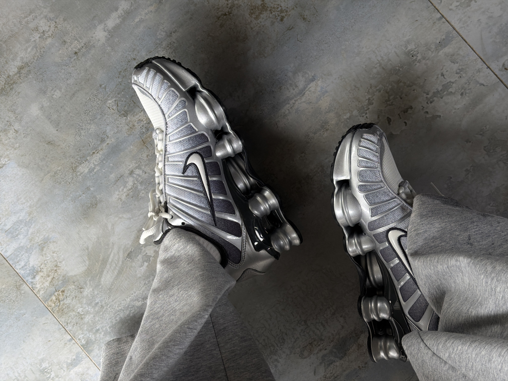 The image shows a person's-eye-view of a pair of silver Nike Shox TL sneakers on a gray concrete floor. The shoes are positioned in a standing stance, partially obscured by gray sweatpants. The setting appears to be an indoor area with a textured concrete floor. No specific indicators suggest Yerevan, Armenia.
