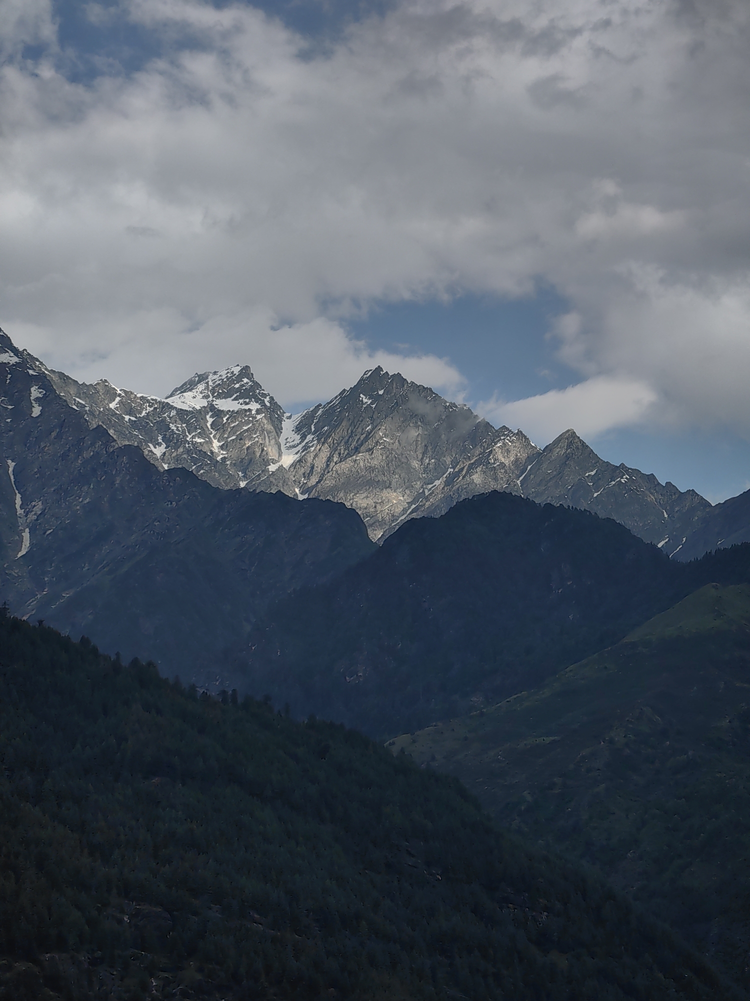 This is a landscape photograph taken in Vashisht, India, featuring a mountain range under a cloudy sky. The foreground consists of dark, forested hills. The midground is dominated by darker, less textured mountains, likely also covered in vegetation. The background showcases a jagged, snow-capped mountain peak, illuminated by sunlight breaking through the clouds. The sky is a mix of grey clouds and patches of blue. No people or specific objects are discernible in the image. The scene depicts a natural, mountainous environment.