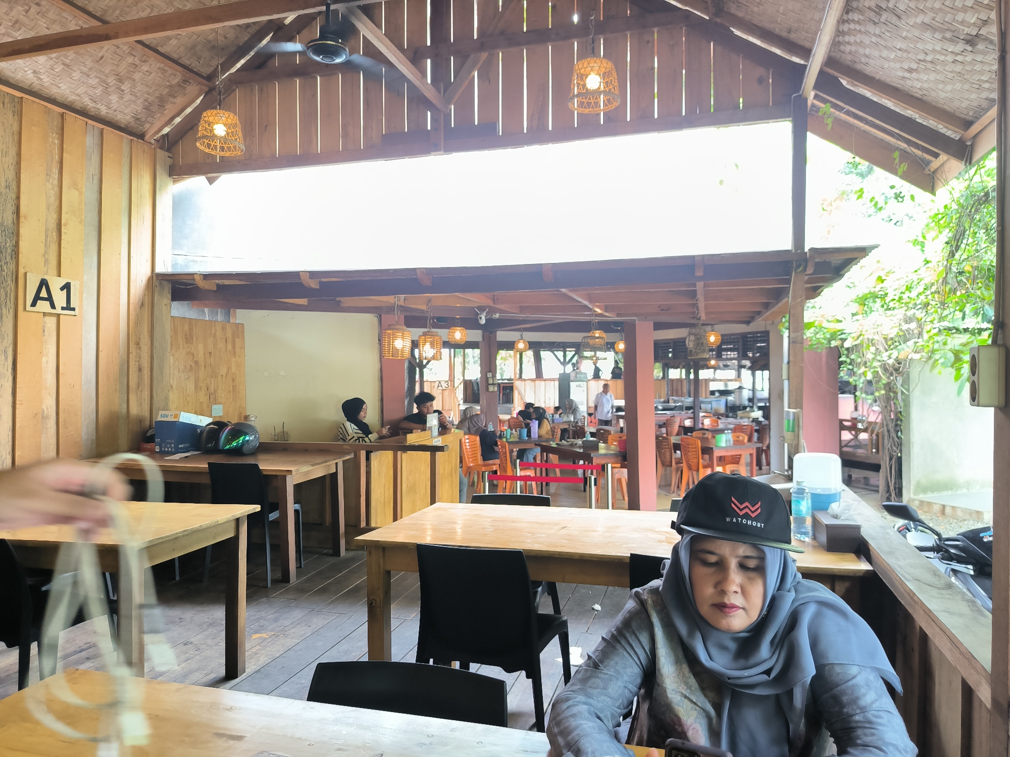 The image depicts an indoor/outdoor dining area with wooden tables and chairs, occupied by multiple individuals. The foreground shows a woman wearing a grey headscarf and a black cap with white lettering, possibly using a mobile device. A motorcycle is parked near her. Other people are seated at tables in the midground. The area is enclosed by wooden structures and features hanging lamps. A sign reading "A1" is visible on a wooden panel. Based on the information provided, the location is Santan, Indonesia.