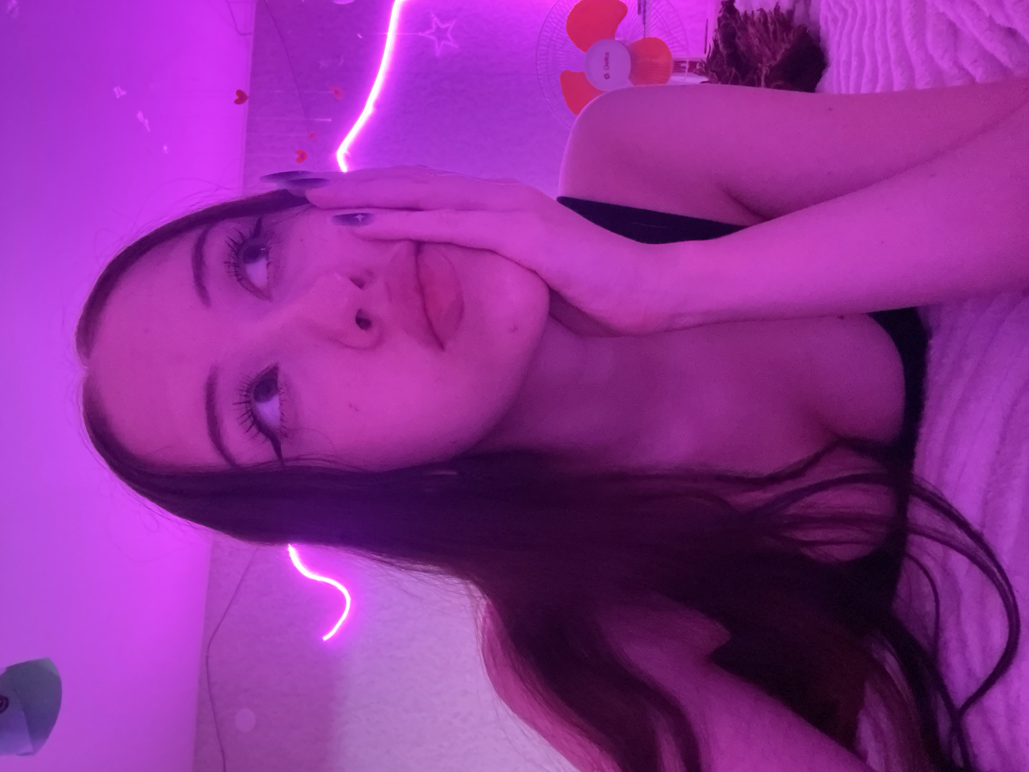 The image shows a young woman posing in a room lit with purple neon lights. She is lying down, with her face close to the camera. Her hand is resting on her cheek, and her eyes are looking upwards. She has long, dark hair that is spread around her. In the background, there are neon lights forming a wavy pattern and a star shape on the wall. A small fan is also visible in the upper right corner of the image. The overall mood is intimate and relaxed, possibly a selfie taken in a personal space. There is no visible text in the image. The context provided indicates the location is Votkinsk, Russia.