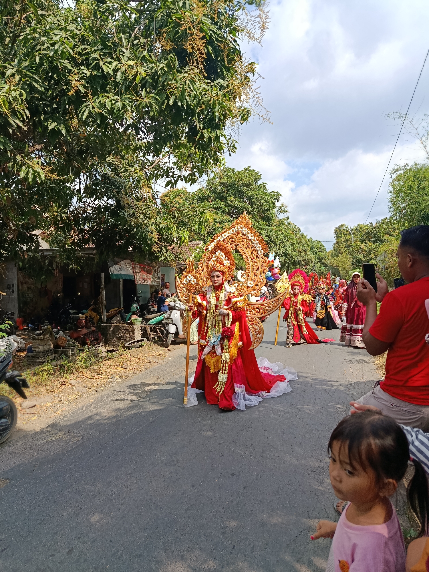 In Ketowan, Indonesia, a colorful parade is underway on a paved road lined with trees and buildings. The central figures are individuals dressed in elaborate, traditional costumes adorned with gold embellishments and red fabrics. One prominent participant in the foreground wears a large, ornate headdress and a flowing red gown, carrying a golden staff. Behind them, other participants in similar festive attire are visible, some appearing to be children. On the right side of the road, a man in a red shirt is taking a photo with his smartphone, while a young child in a pink shirt looks on. The environment appears to be a street scene with parked motorcycles and roadside stalls, suggesting a community event. The sky is partly cloudy, indicating an outdoor daytime setting.