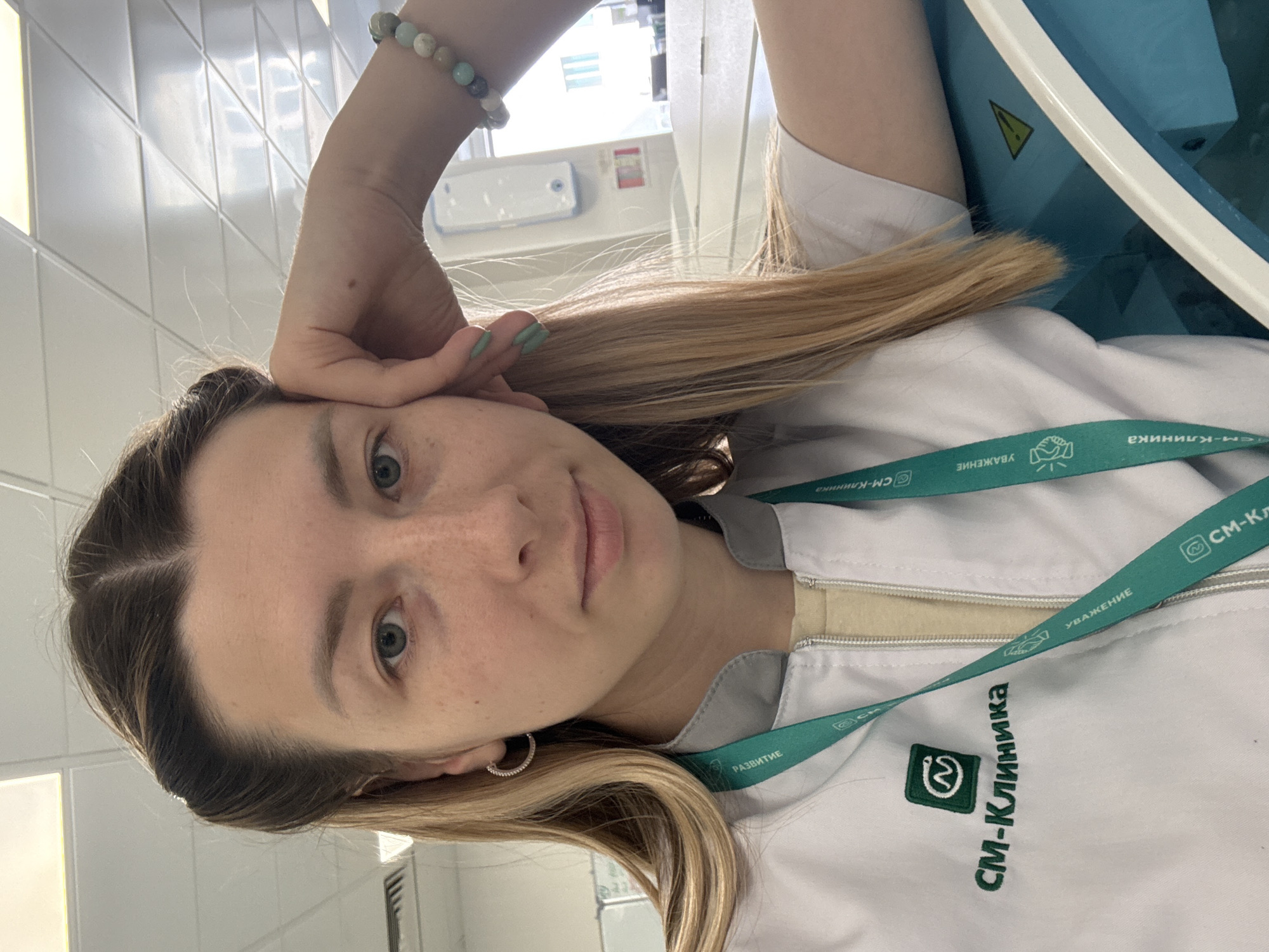 A fair-skinned woman with light brown hair and freckles is posing indoors. Her hair is parted in the middle and styled straight. She has her hand on her forehead, and is wearing a beaded bracelet on her wrist. She is wearing a white lab coat or medical jacket over a light yellow shirt. She is wearing a green lanyard with the logo "СМ-Клиника" (SM-Clinic) on it, which is a chain of medical clinics in Moscow, Russia. The background is a sterile environment with white tiled walls. There is a piece of medical equipment visible in the corner.